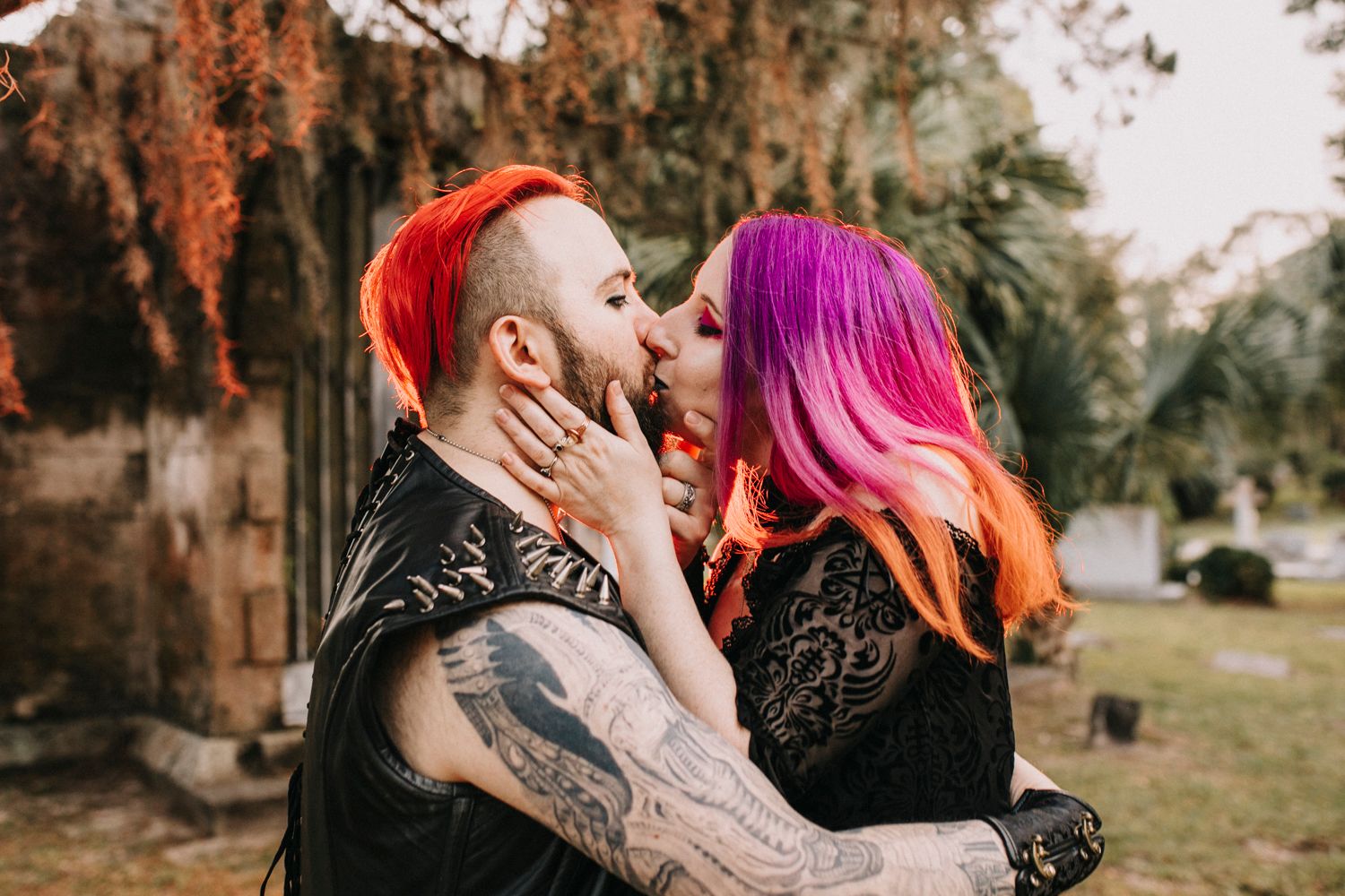 Colorful couple with dyed hair sharing a romantic kiss outdoors among autumn foliage in a gothic-inspired setting.