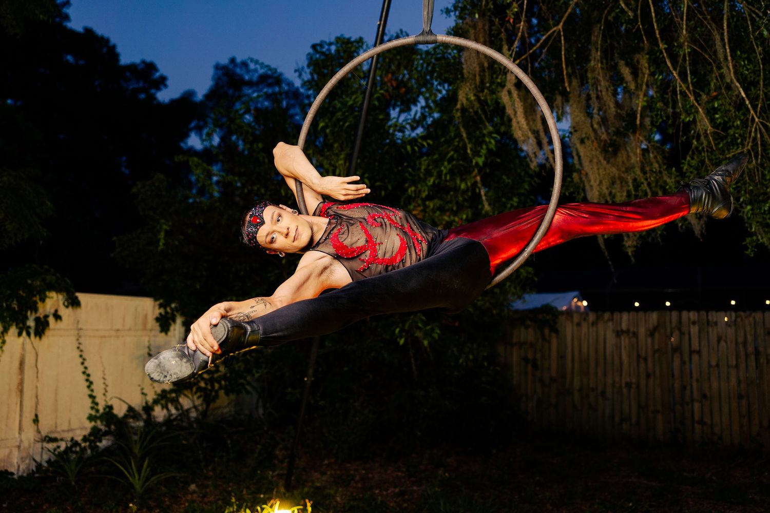 Aerial hoop performer in red costume gracefully posing on a lyra ring suspended outdoors during evening twilight.