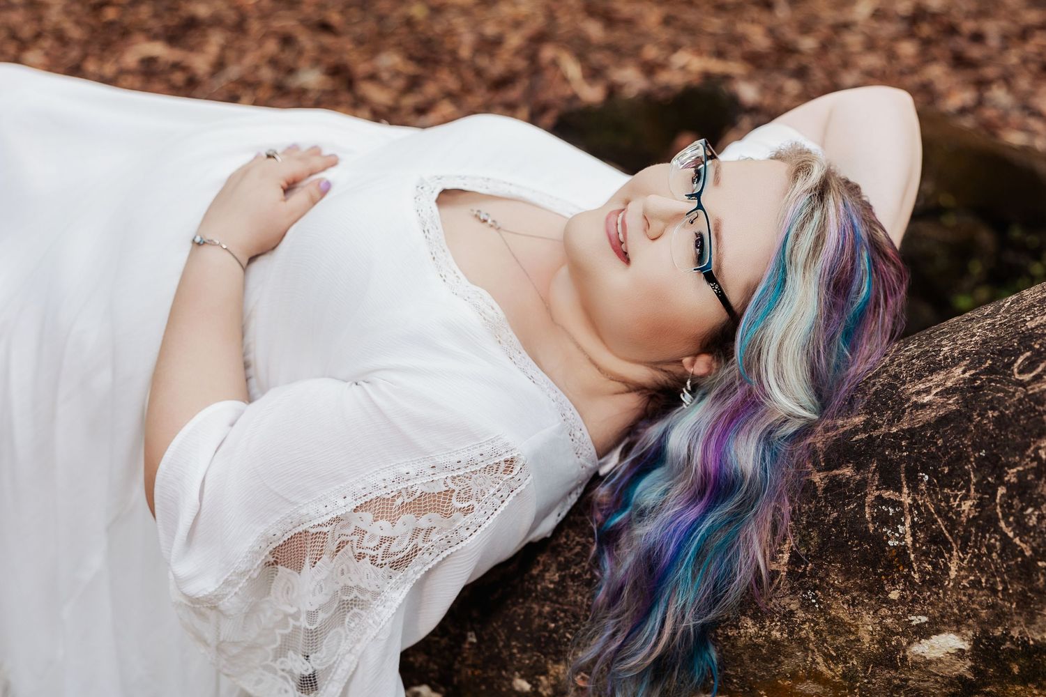Woman with colorful rainbow hair wearing white lace dress, reclining on rocks outdoors in autumn setting.