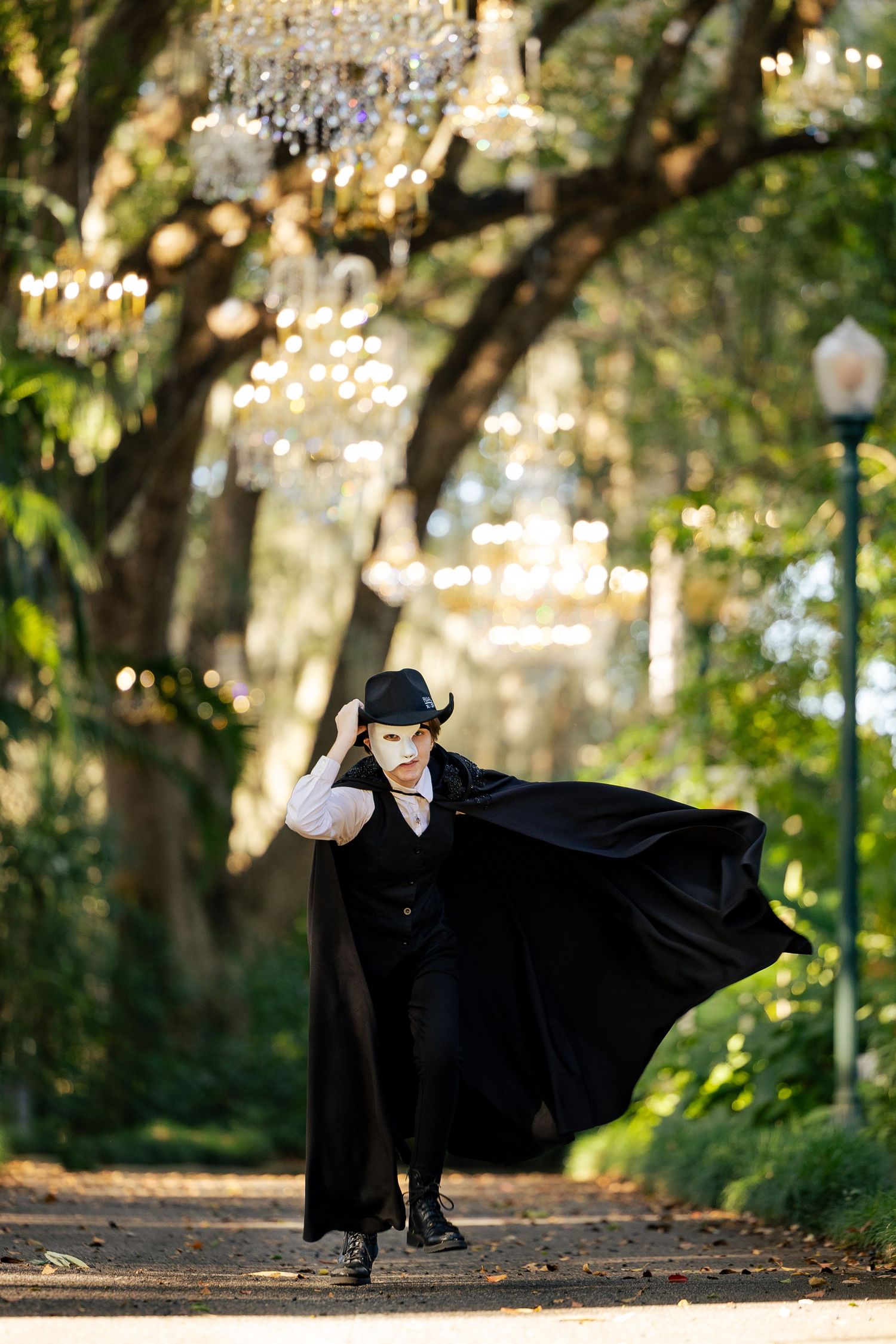 Person dressed as Phantom of Opera in black cape and mask walks along chandelier-lined outdoor path.