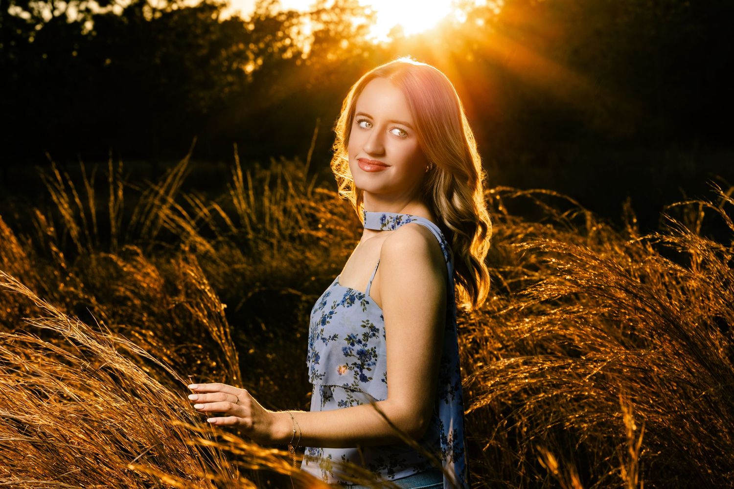 Young woman in floral dress standing in golden sunset light among tall grasses.