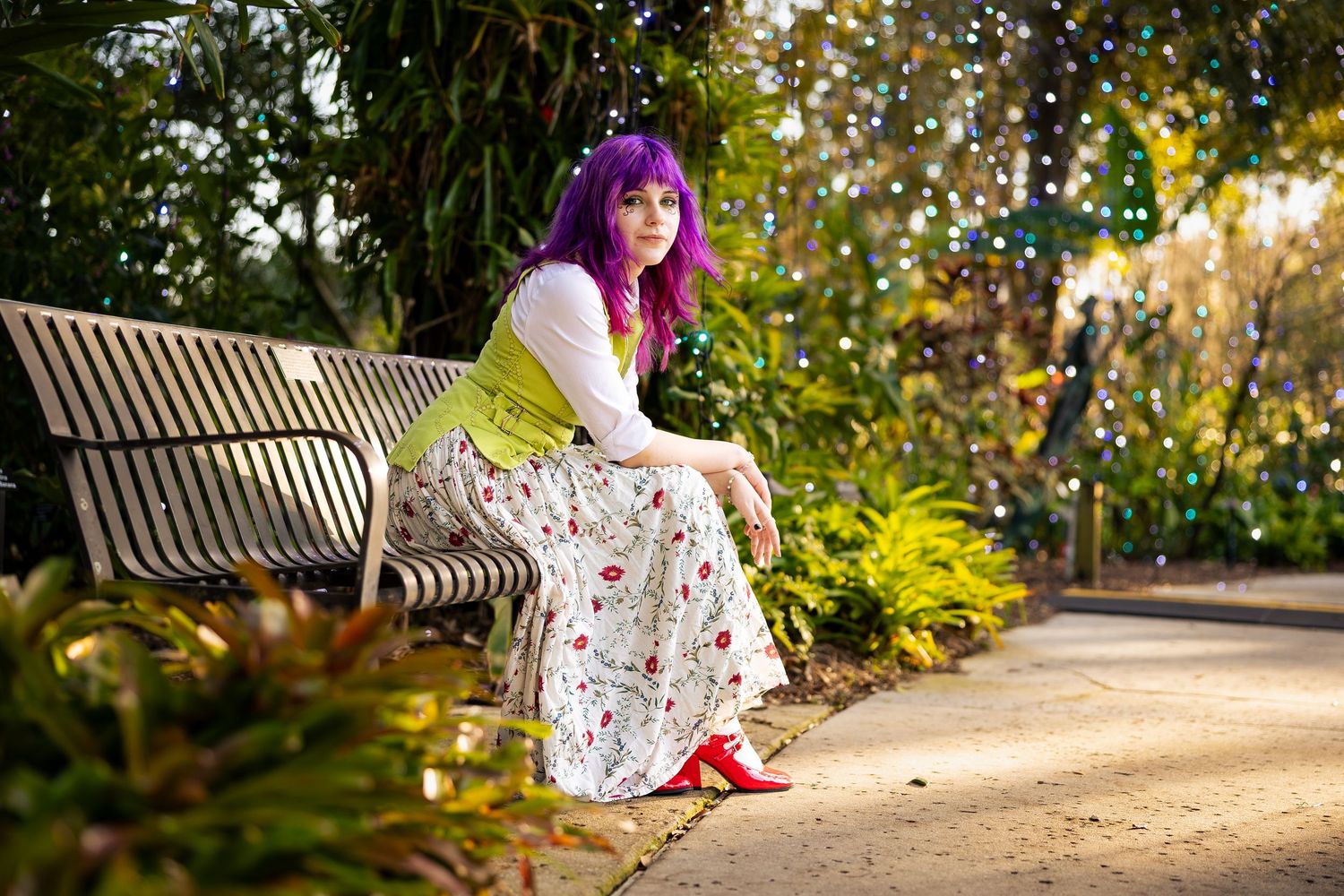 Woman with purple hair in floral skirt sitting on park bench surrounded by lush green gardens.