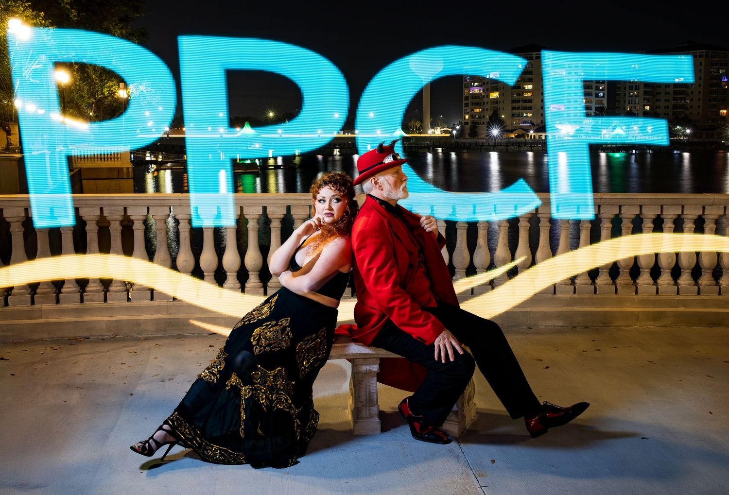 Couple in formal attire poses back-to-back at night with glowing PPCF letters over waterfront.