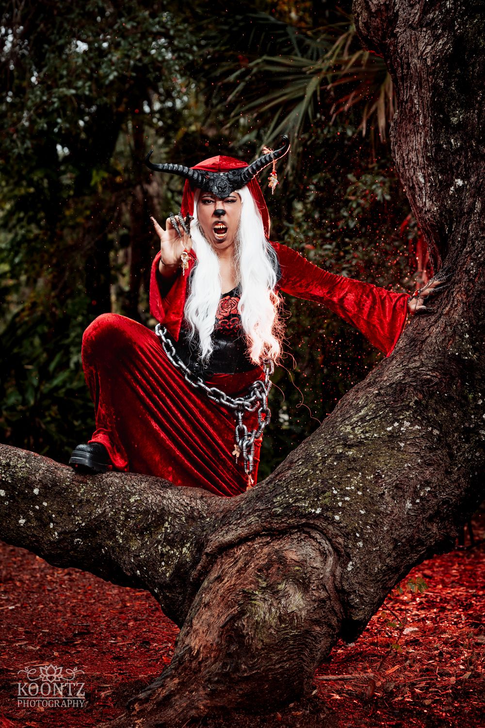 A cosplayer in a red velvet suit with black horns and chains sits dramatically in a large mossy tree outdoors.
