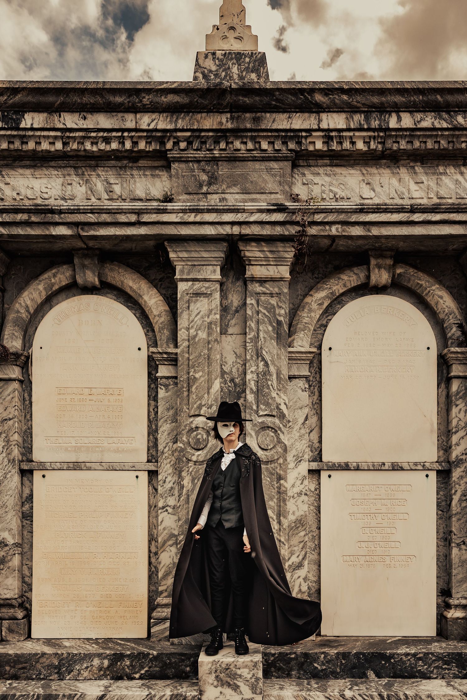 Gothic caped figure in top hat standing before grand marble mausoleum facade under dramatic sky.