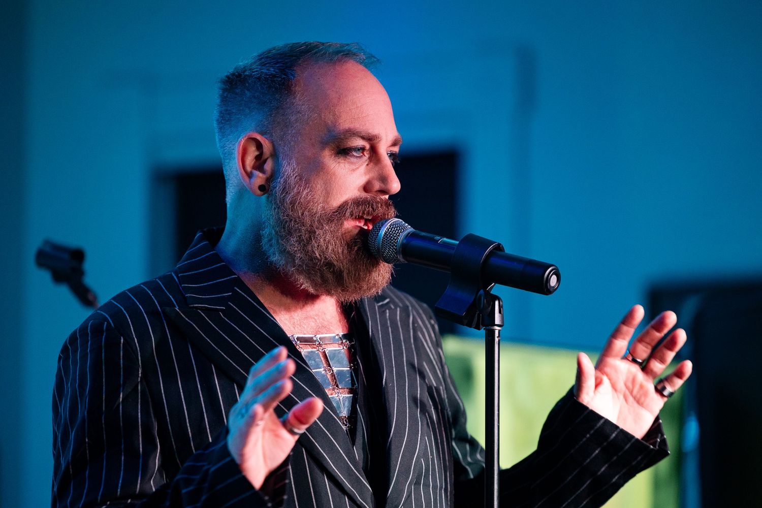 Bearded male singer performing on stage, wearing a pinstripe suit, speaking into a microphone under blue stage lighting.