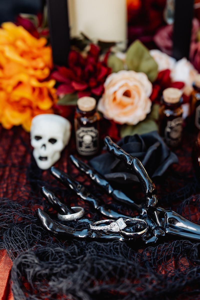 Halloween-themed wedding rings displayed on a black skeleton hand with skulls and flowers decor.