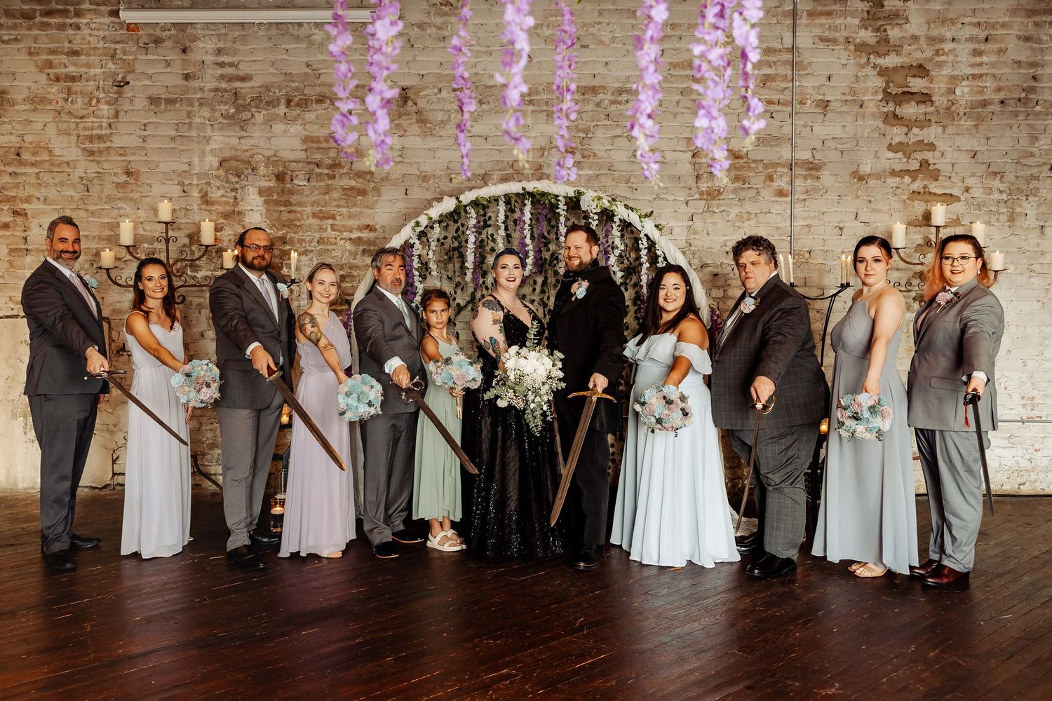 Wedding party poses with swords before circular floral arch with purple wisteria in brick venue.