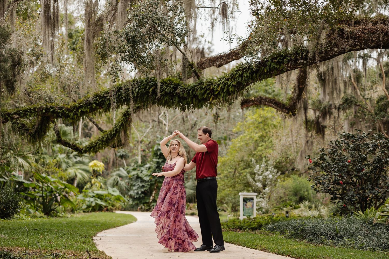 Couple dancing elegantly on a garden path beneath sprawling moss-covered oak trees.