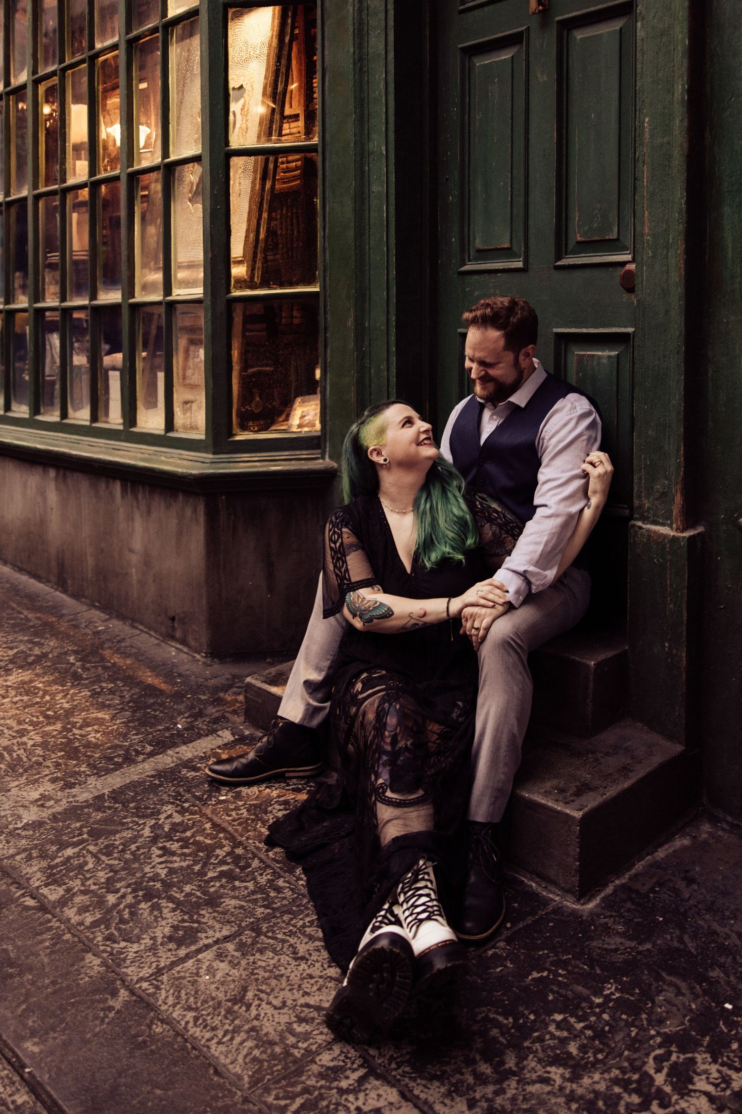 A romantic couple sits on steps outside a vintage storefront with glowing windows at dusk.
