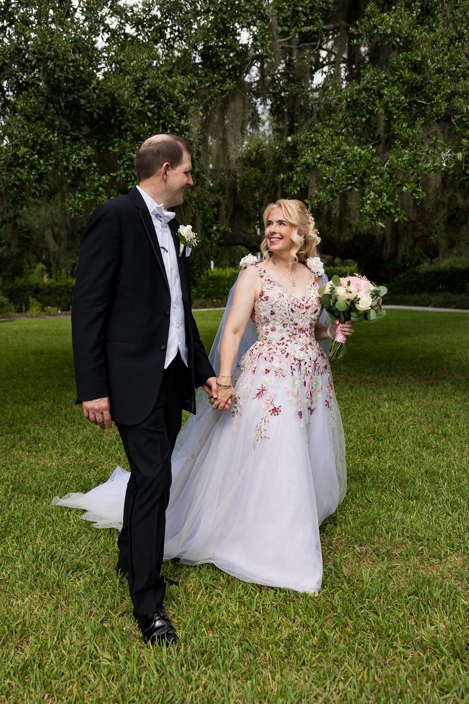 Newlyweds walking hand in hand through lush green lawn beneath canopy of Spanish moss trees.