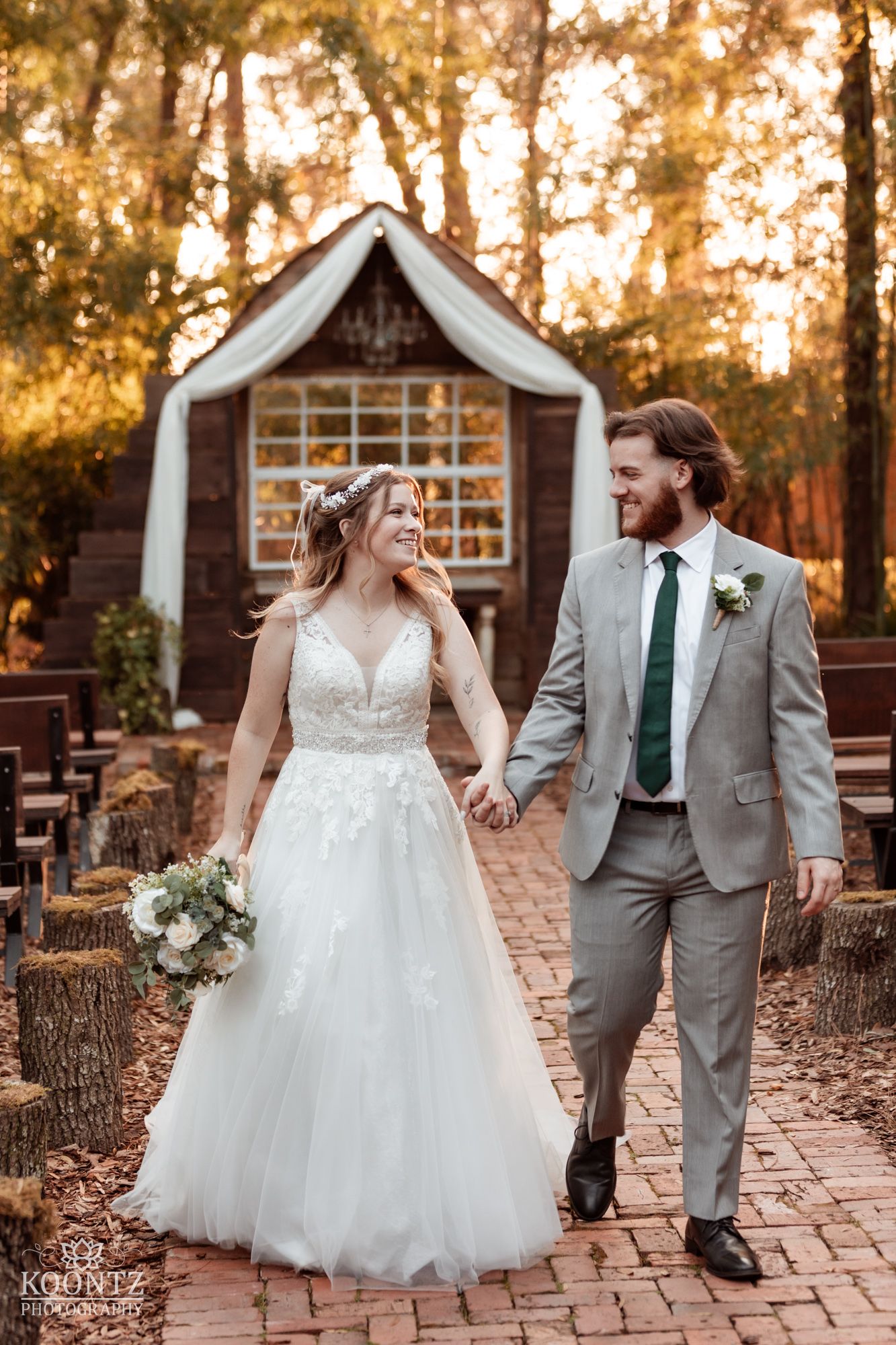 Bride and groom walk hand in hand down brick aisle at rustic outdoor wedding venue surrounded by tall trees.
