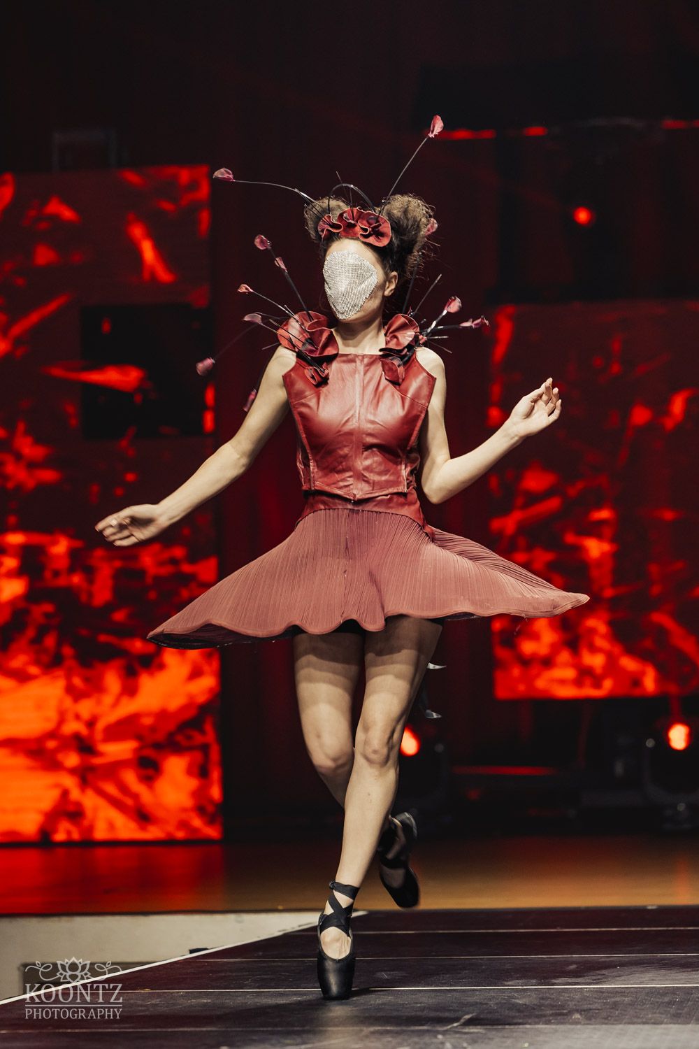 Dancer in a pink dress performs an energetic leap on stage with red lighting effects.