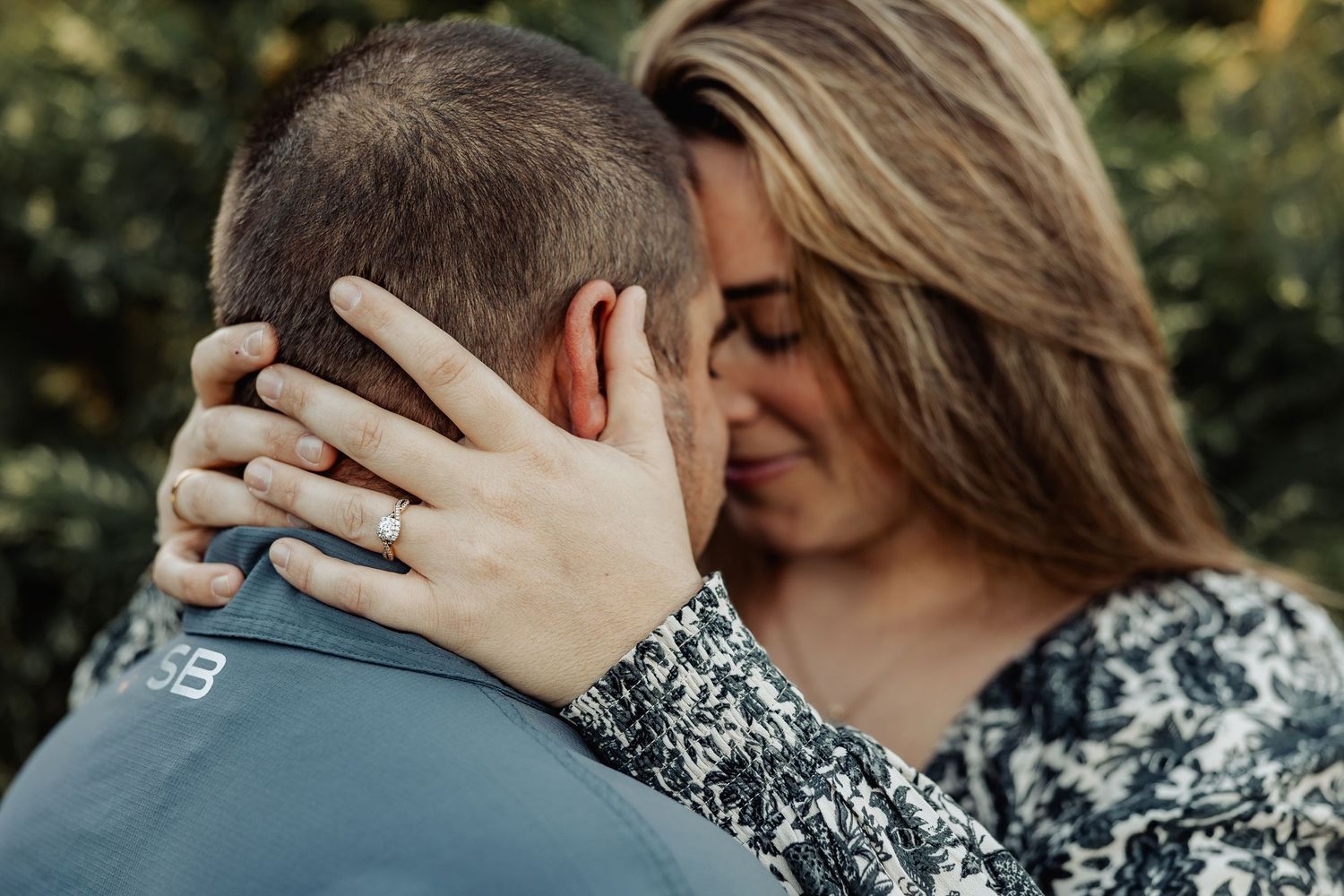 A woman with an engagement ring tenderly embraces her partner outdoors among green foliage.