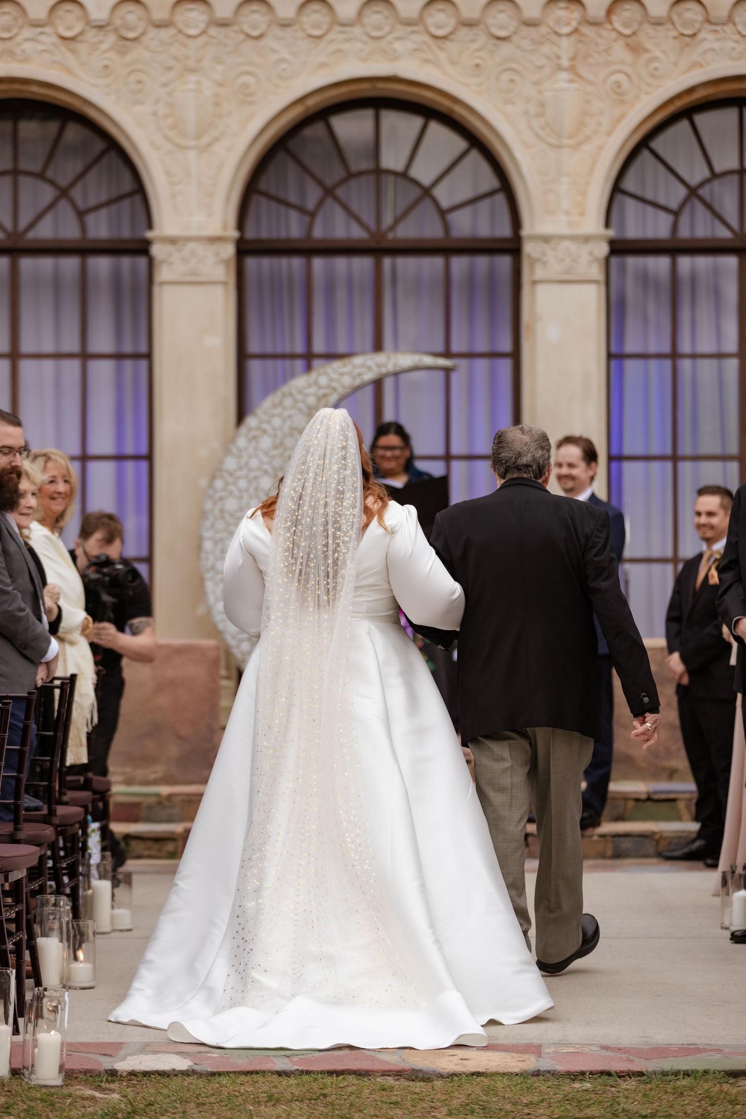 Bride in white gown and veil walks down aisle with escort toward crescent moon altar backdrop.