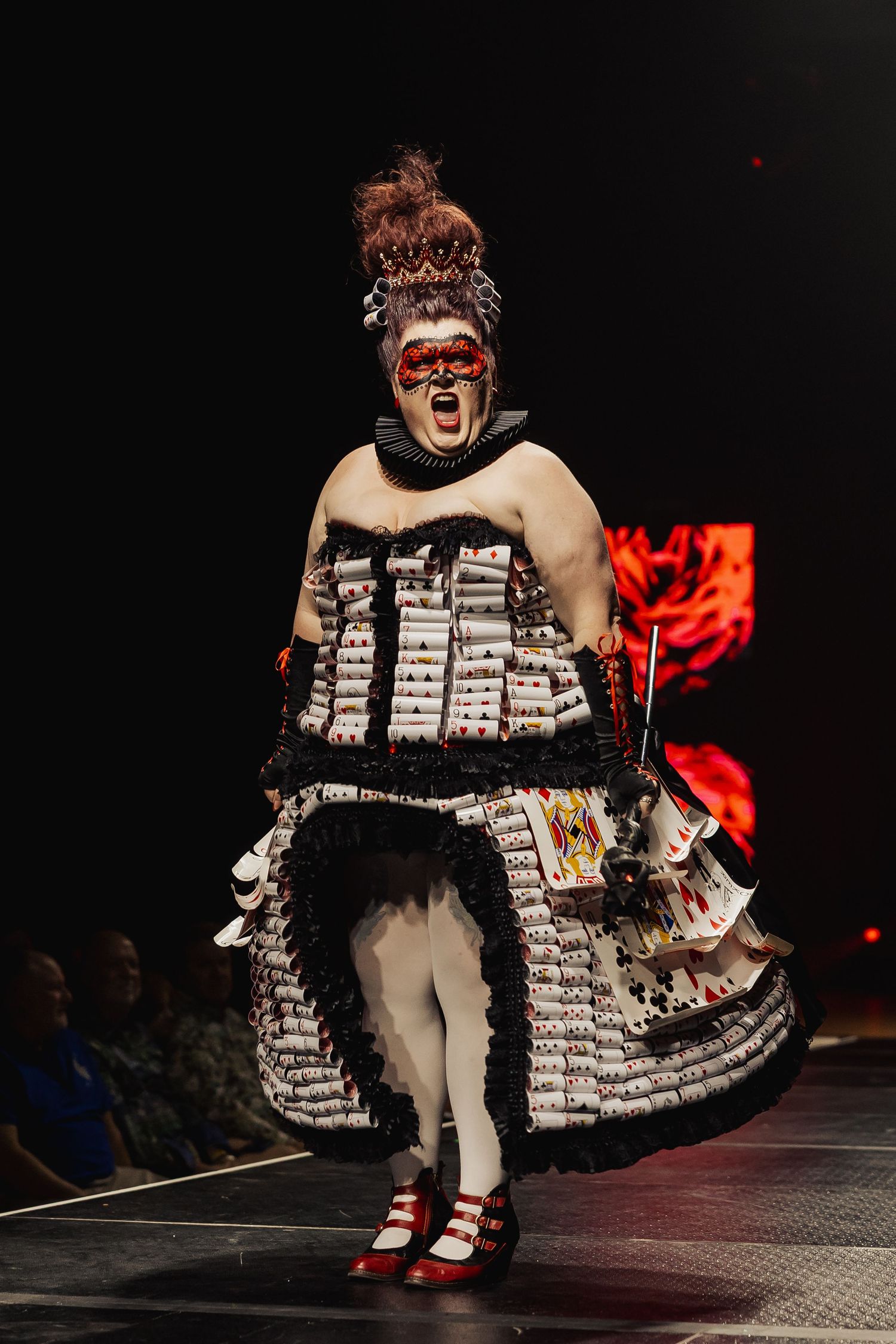 Performer in dramatic playing card costume with red mask walks runway at theatrical fashion show.