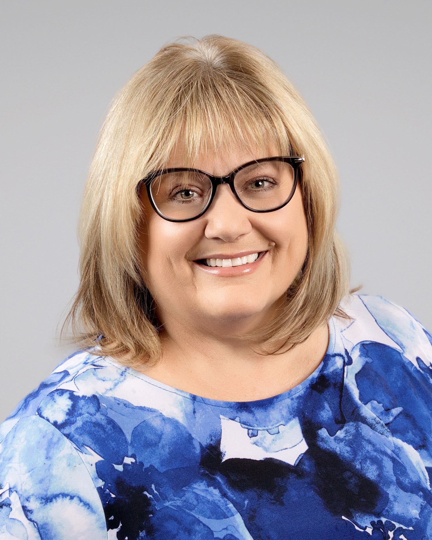 Smiling blonde woman with glasses wearing a blue floral top in a professional headshot.