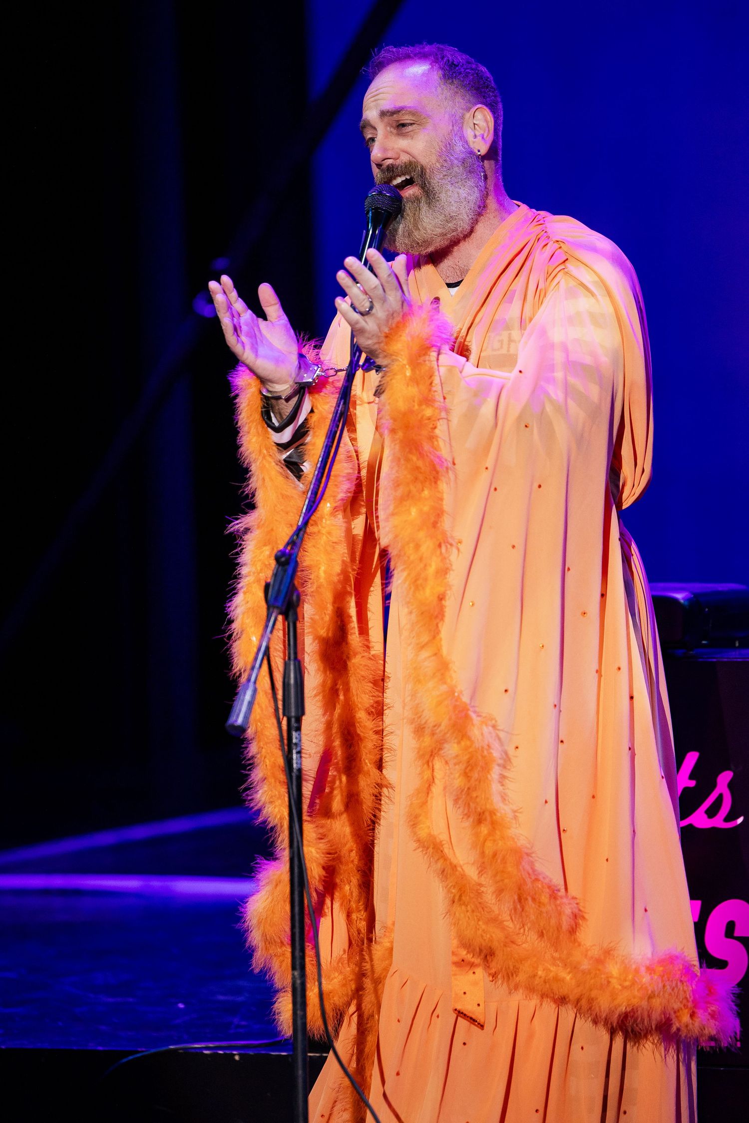 A male performer in an orange feather-trimmed robe sings into a microphone on a purple-lit cabaret stage.
