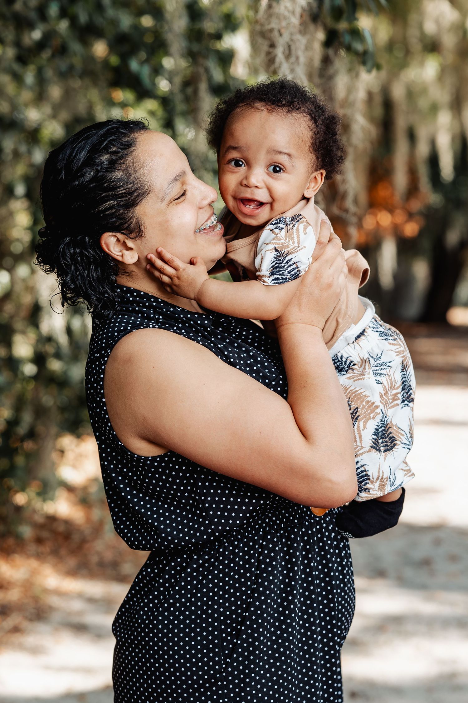 A woman joyfully holds a smiling baby boy outdoors among lush green trees.