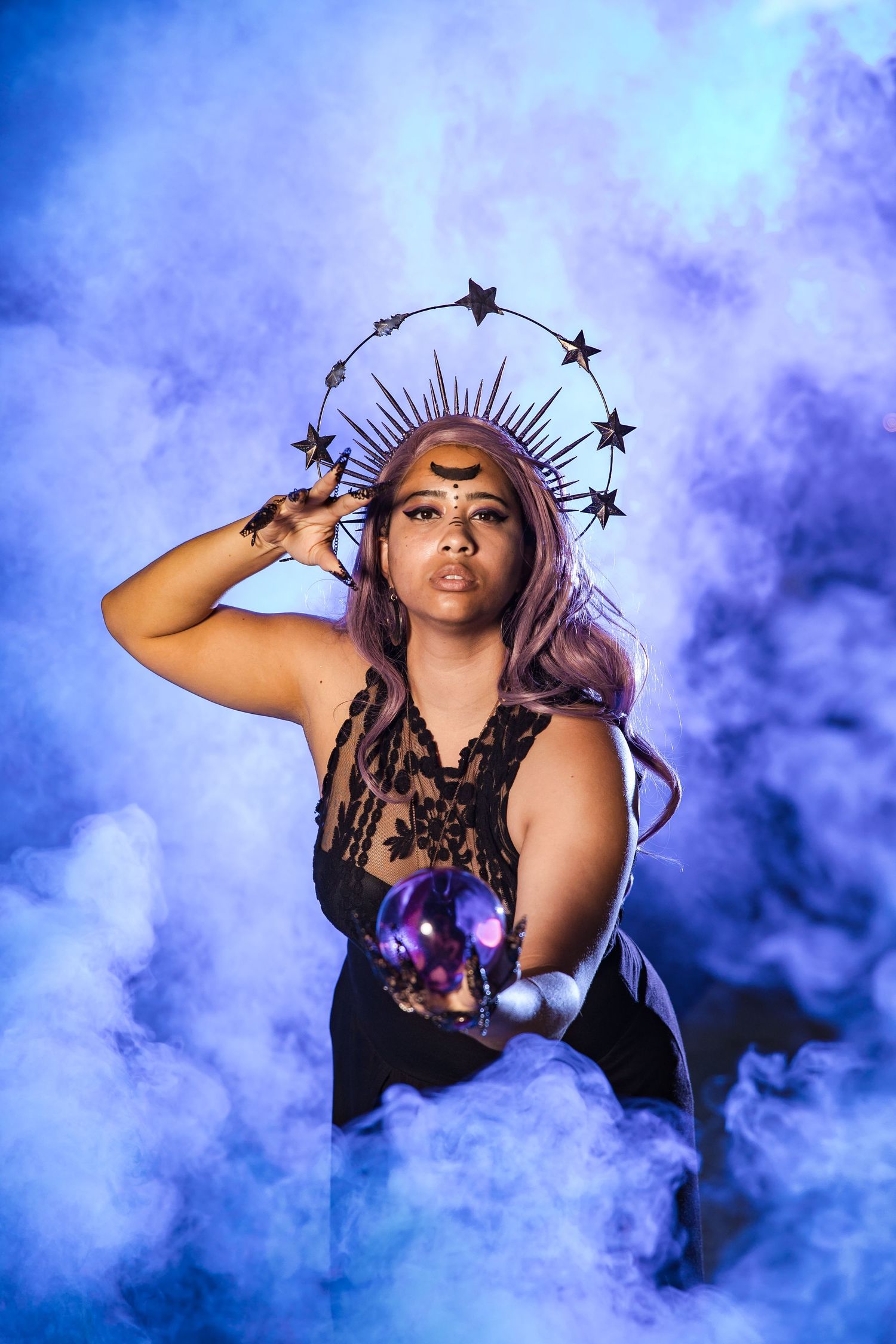 A mystical fortune teller in black lace holds a crystal ball amid blue smoke, wearing a star crown and purple hair.