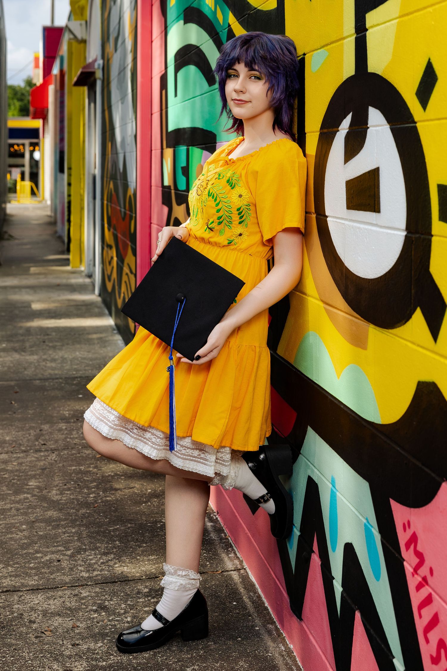 Young woman in yellow dress holding graduation cap, posing against colorful graffiti mural wall.