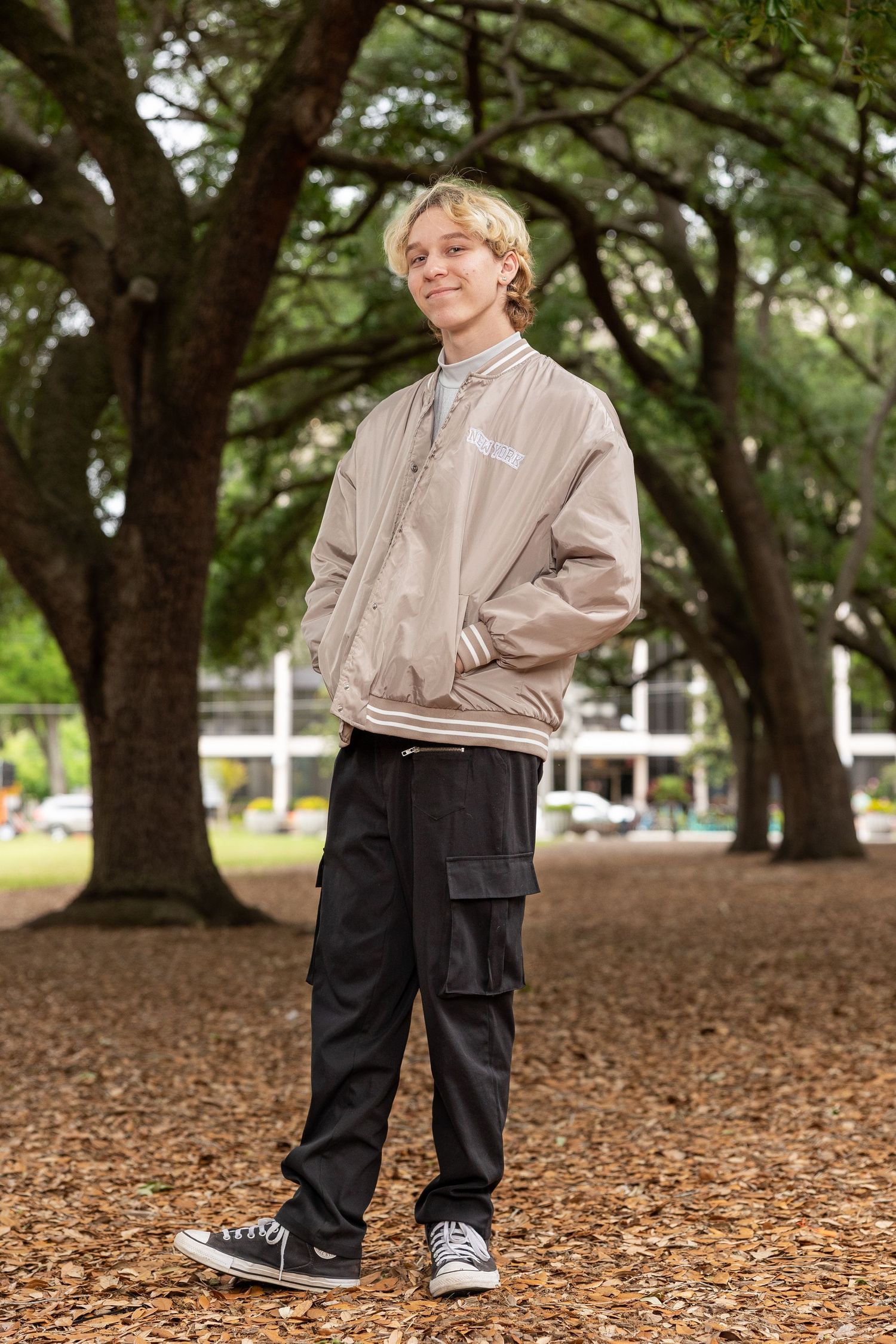 Teen boy in beige bomber jacket and black cargo pants standing in a tree-lined park smiling.