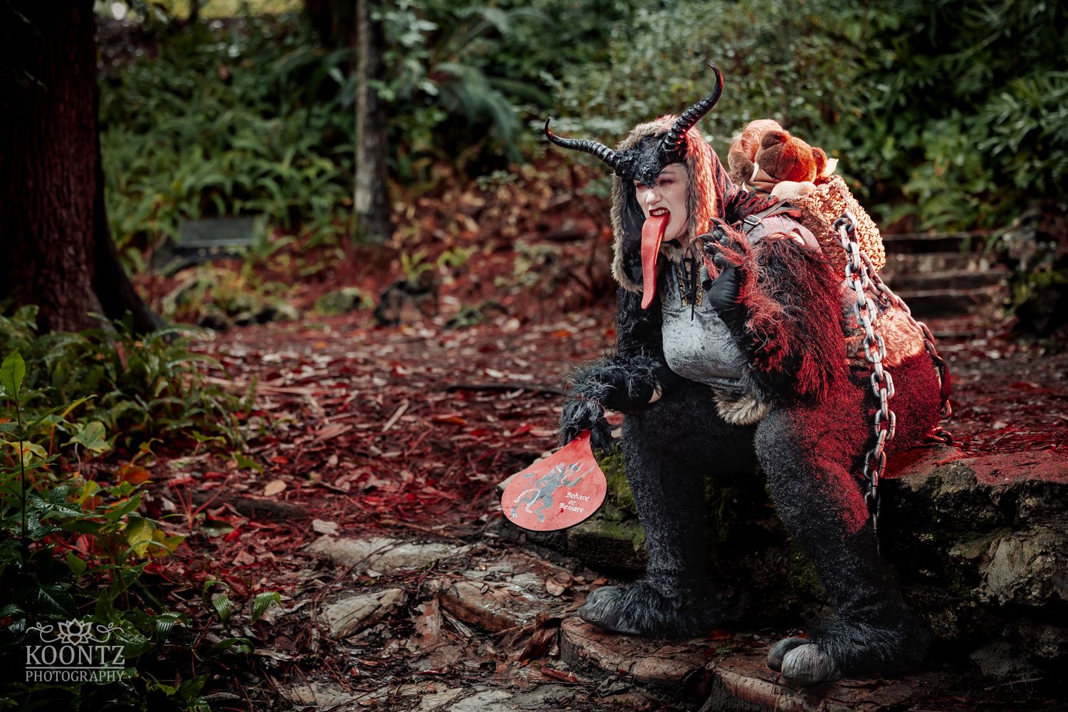 Person in elaborate dark creature costume crouches on forest ground surrounded by red autumn foliage.