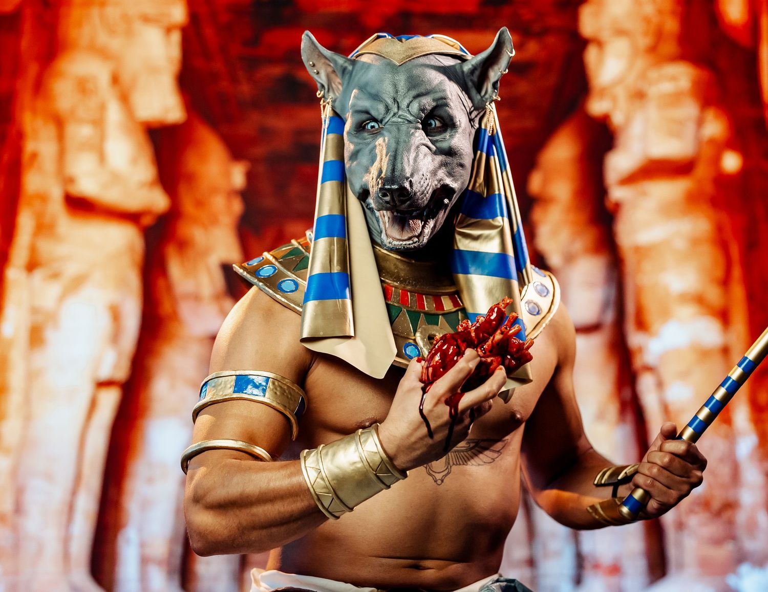 Performer in Anubis Egyptian god costume with jackal mask holding red scepter in ancient temple.