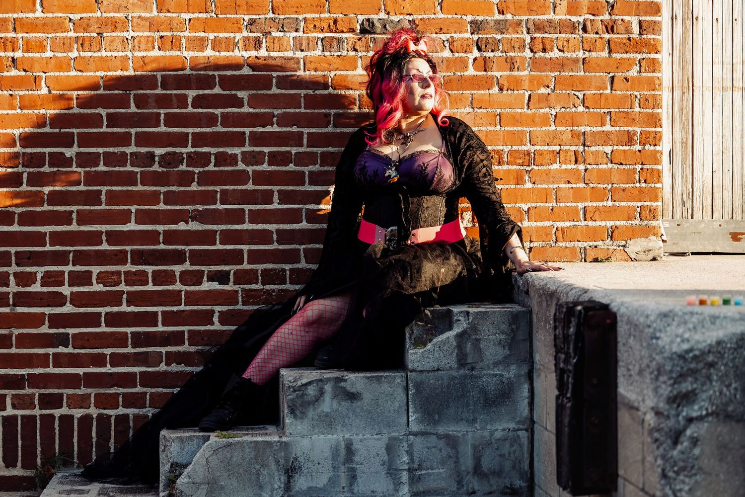 Woman in gothic purple and black costume sits on stone steps against a sunlit brick wall.