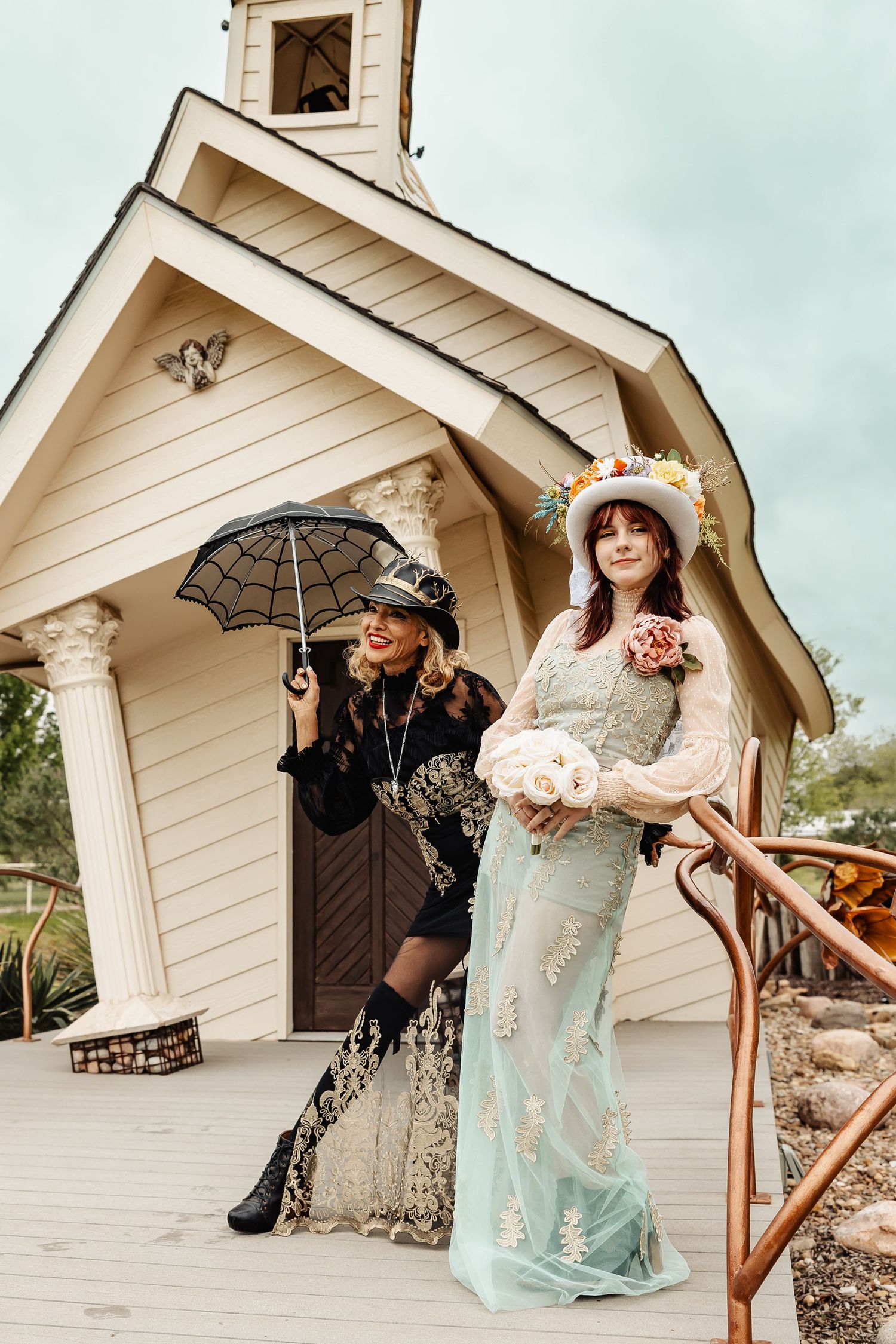 Two women in vintage-inspired outfits pose outside white chapel, one in mint bridal gown, one with spider web umbrella.