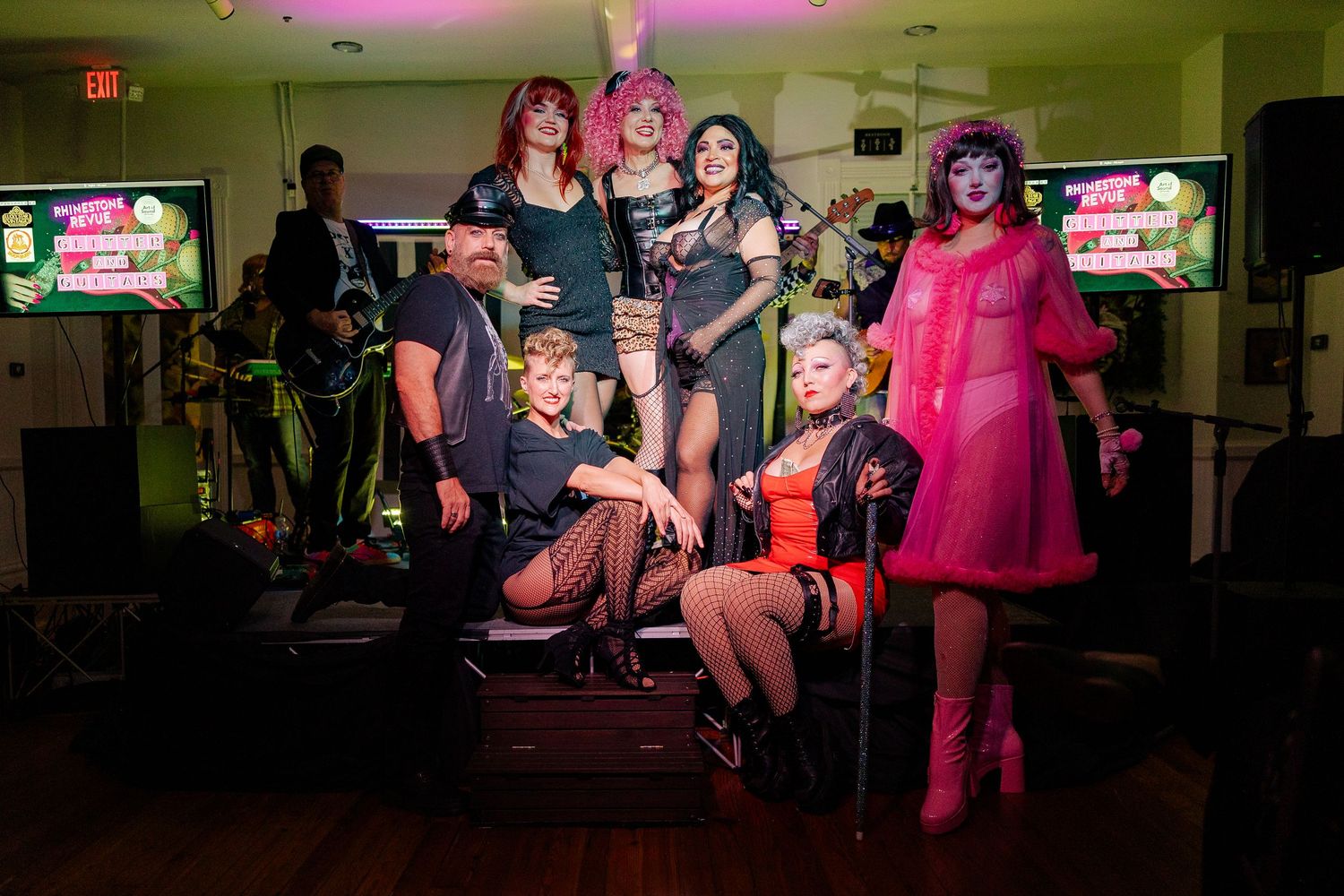 Colorful drag performers and band members pose together at a vibrant nightclub event with pink and green lighting.