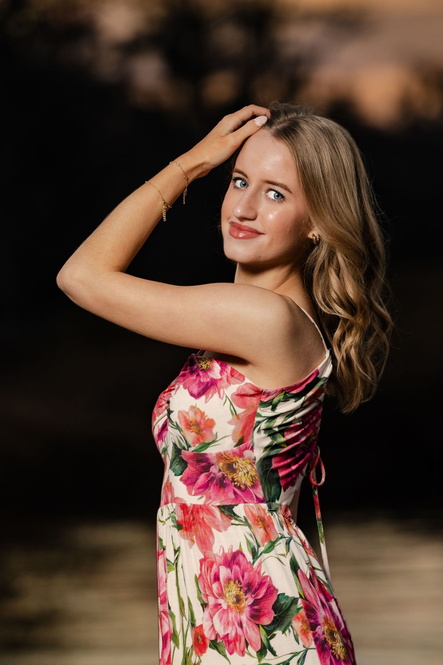 Young woman in floral pink dress poses outdoors at sunset, hand in blonde hair, smiling softly.