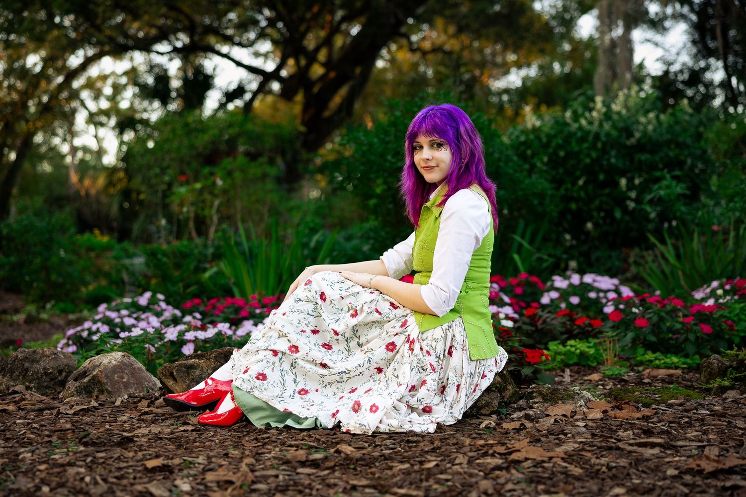Woman with purple hair in floral dress and red shoes sitting in a garden with pink flowers.