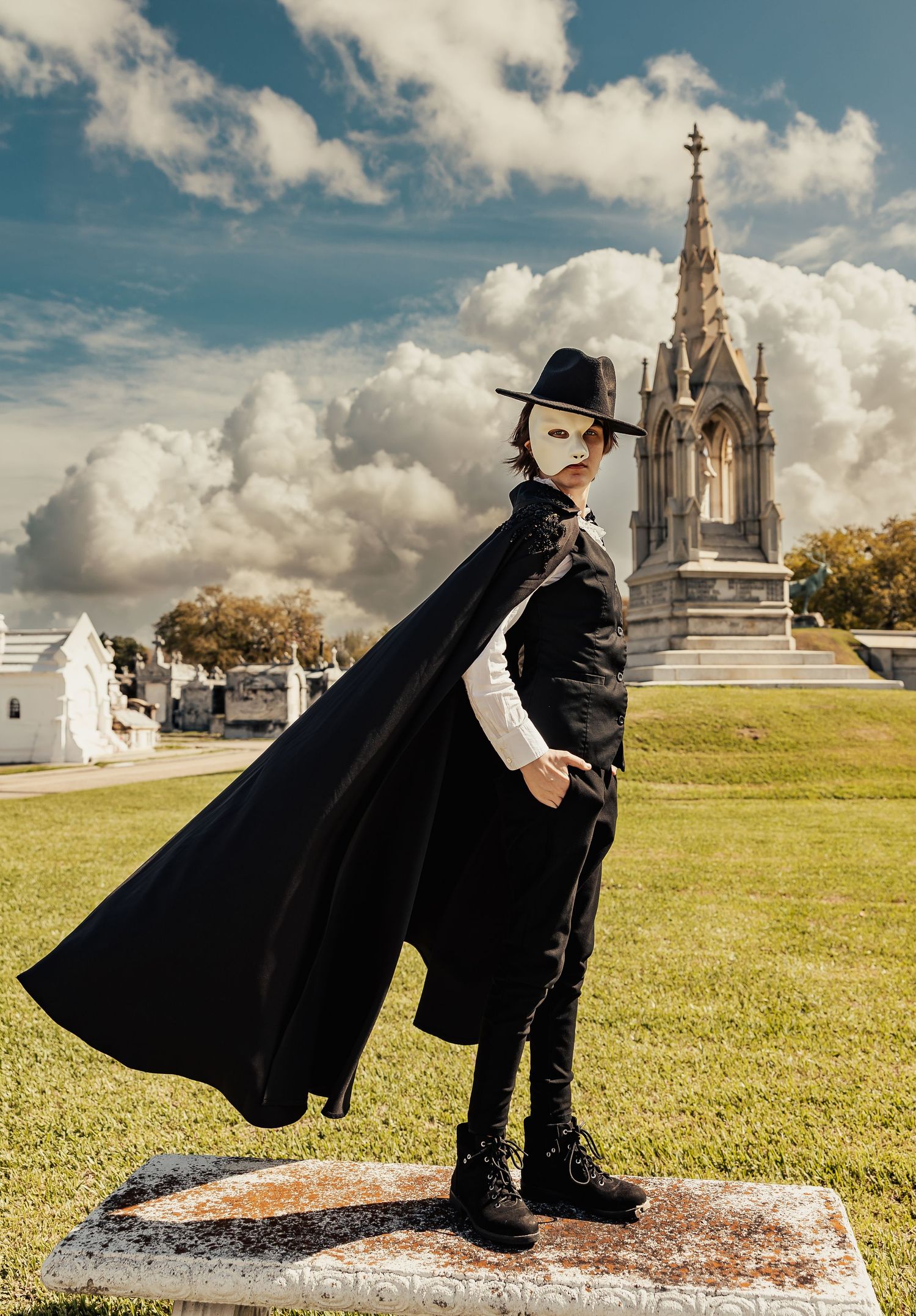 Phantom of the Opera costumed figure in white mask and black cape poses in sunlit cemetery.