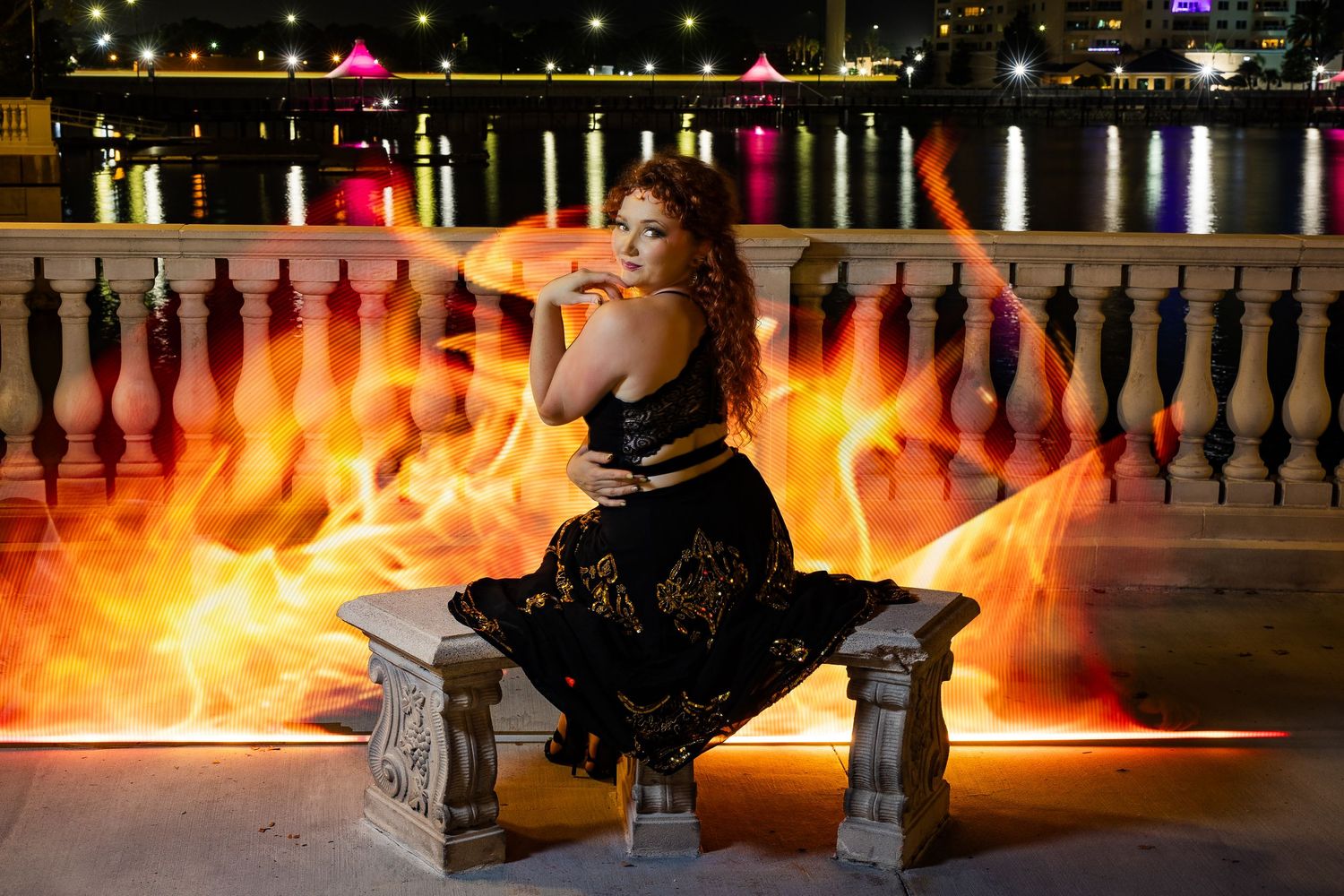 Woman in black gold dress sits on bench surrounded by dramatic fire effects at night.