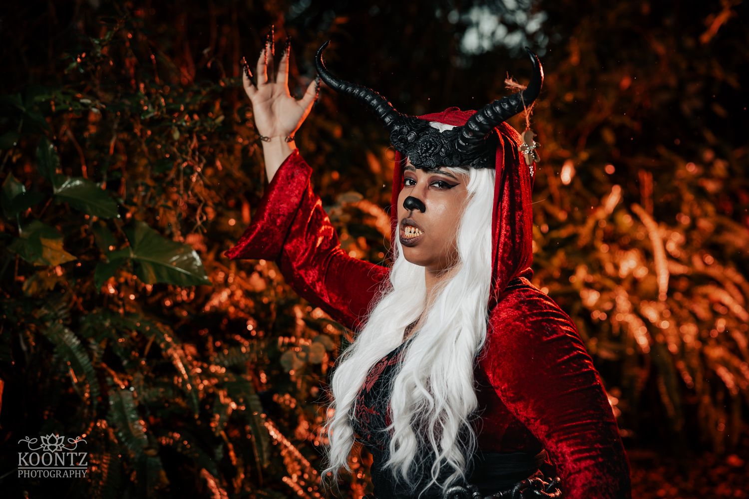 Person in devil cosplay with black horns and white wig poses dramatically among red autumn foliage.