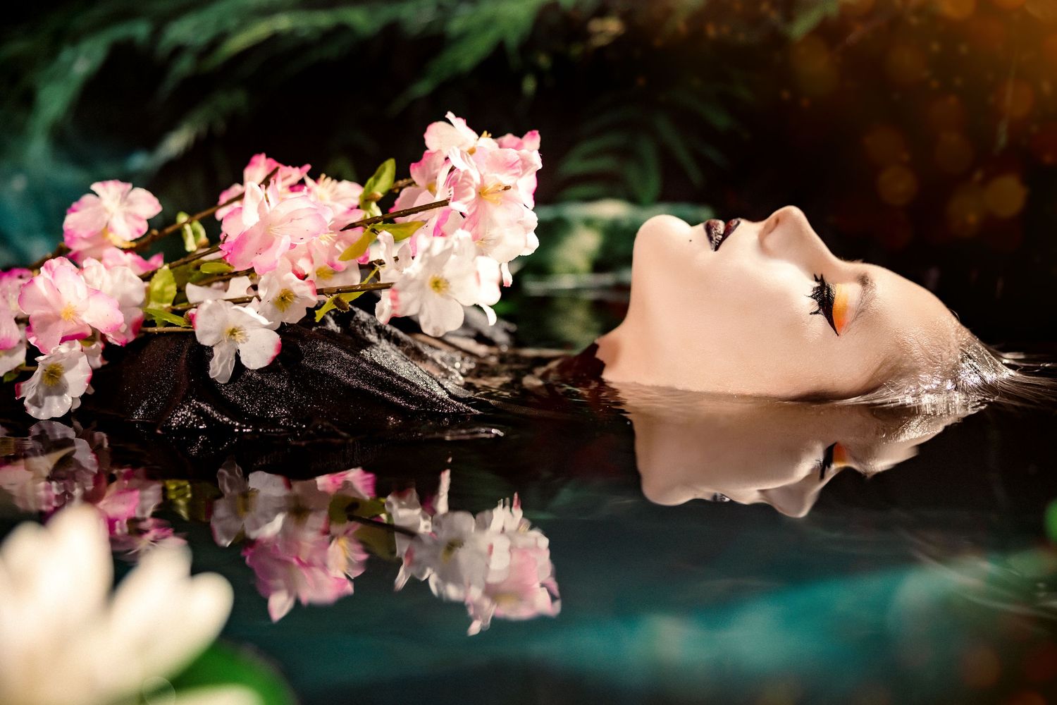 Serene woman floating in dark reflective water surrounded by pink cherry blossoms.