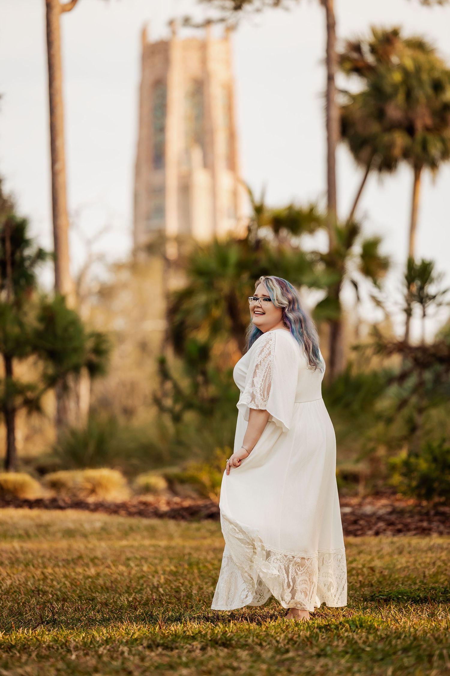 Woman in white lace dress smiling in park with palm trees and tower in background.
