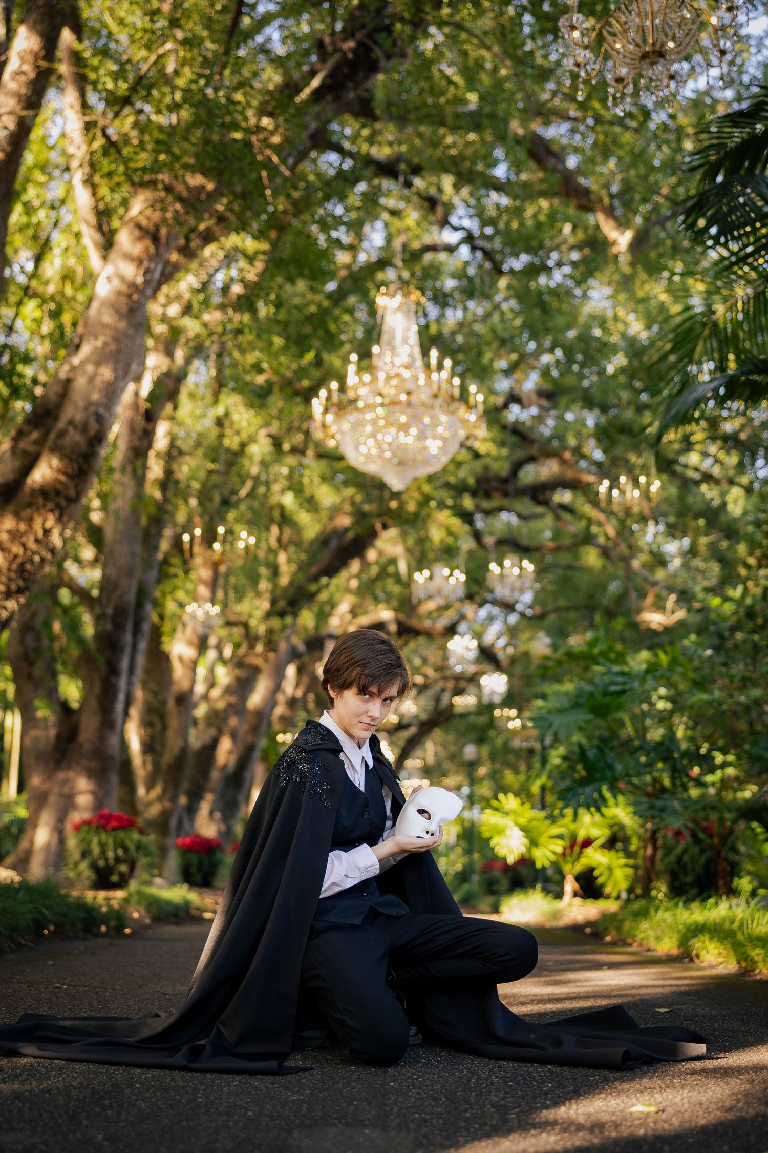 Person in black cape costume kneeling outdoors holding white mask, crystal chandelier hanging among lush green trees.