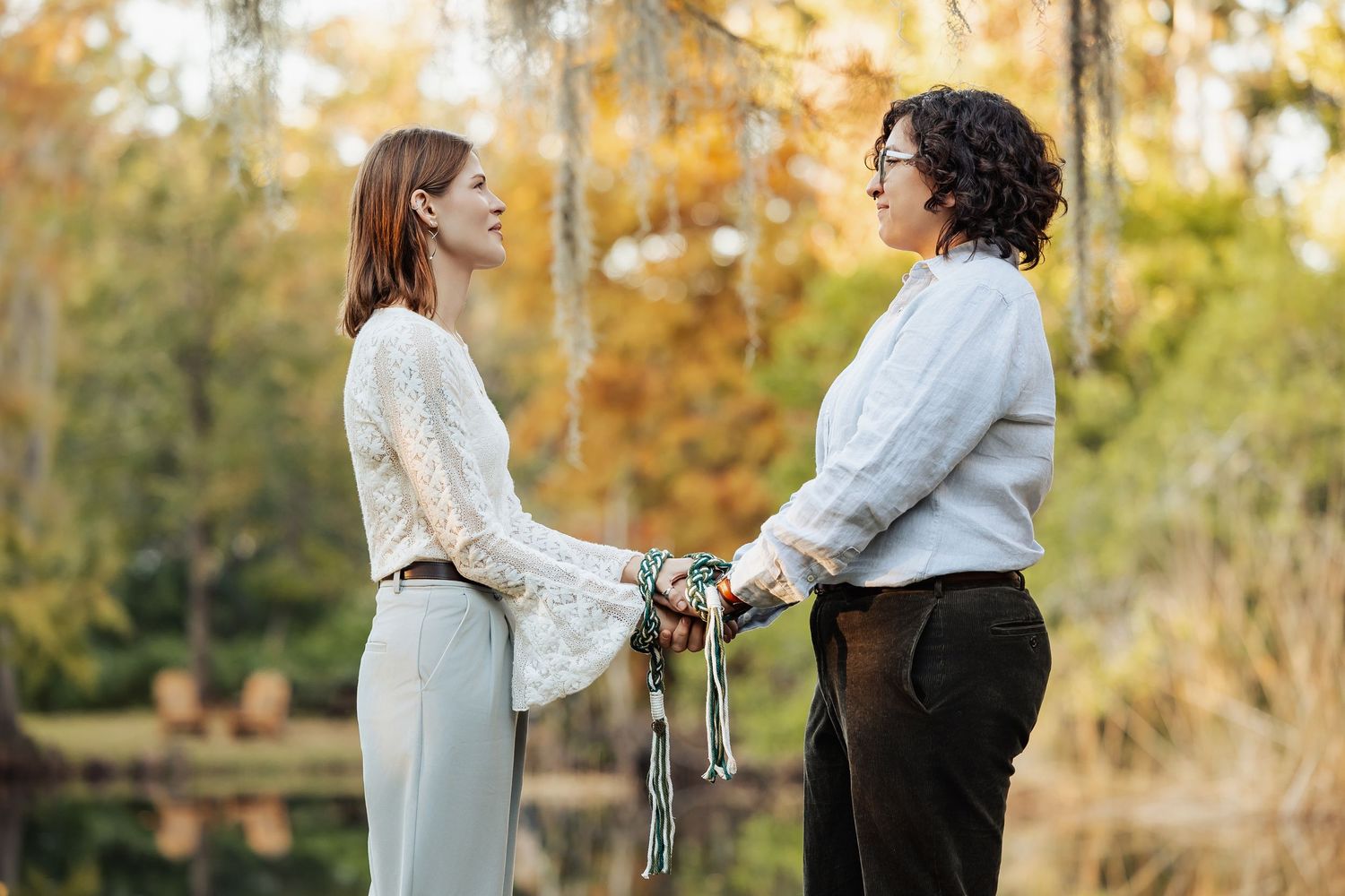 Couple holds hands during an intimate outdoor handfasting ceremony surrounded by autumn foliage.