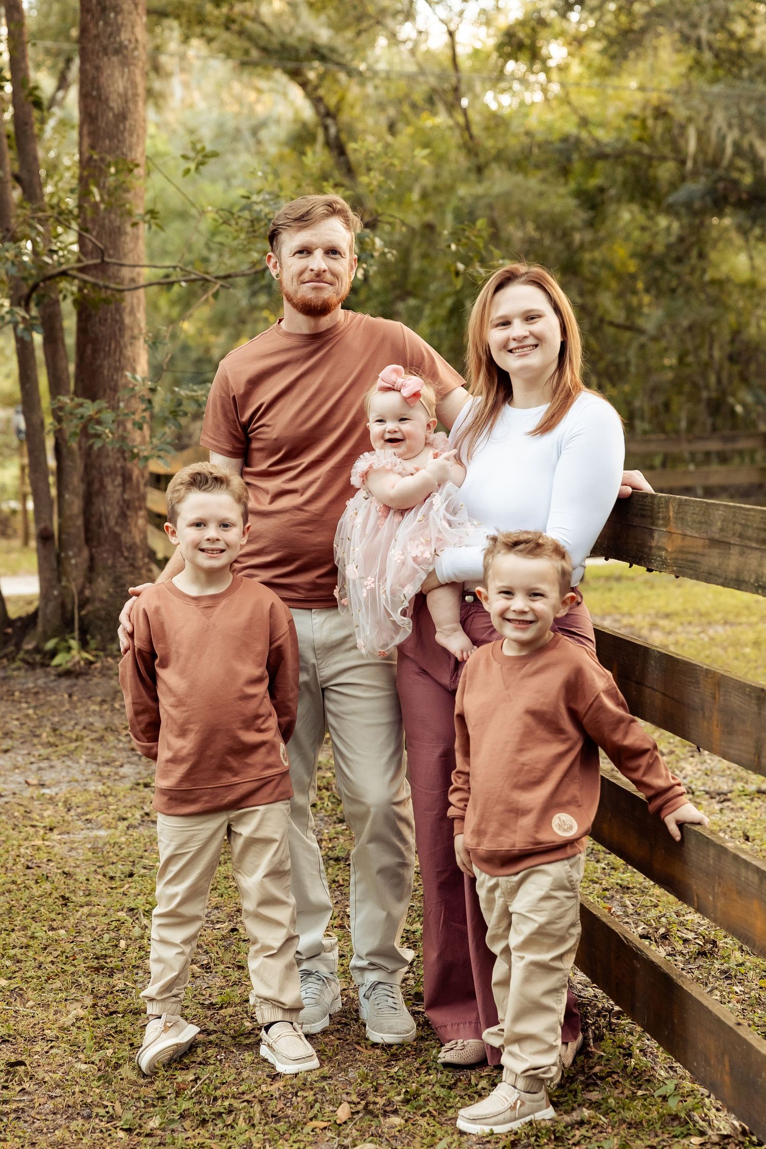 Family of five in coordinating earth tones poses together by a wooden fence in a forest park.
