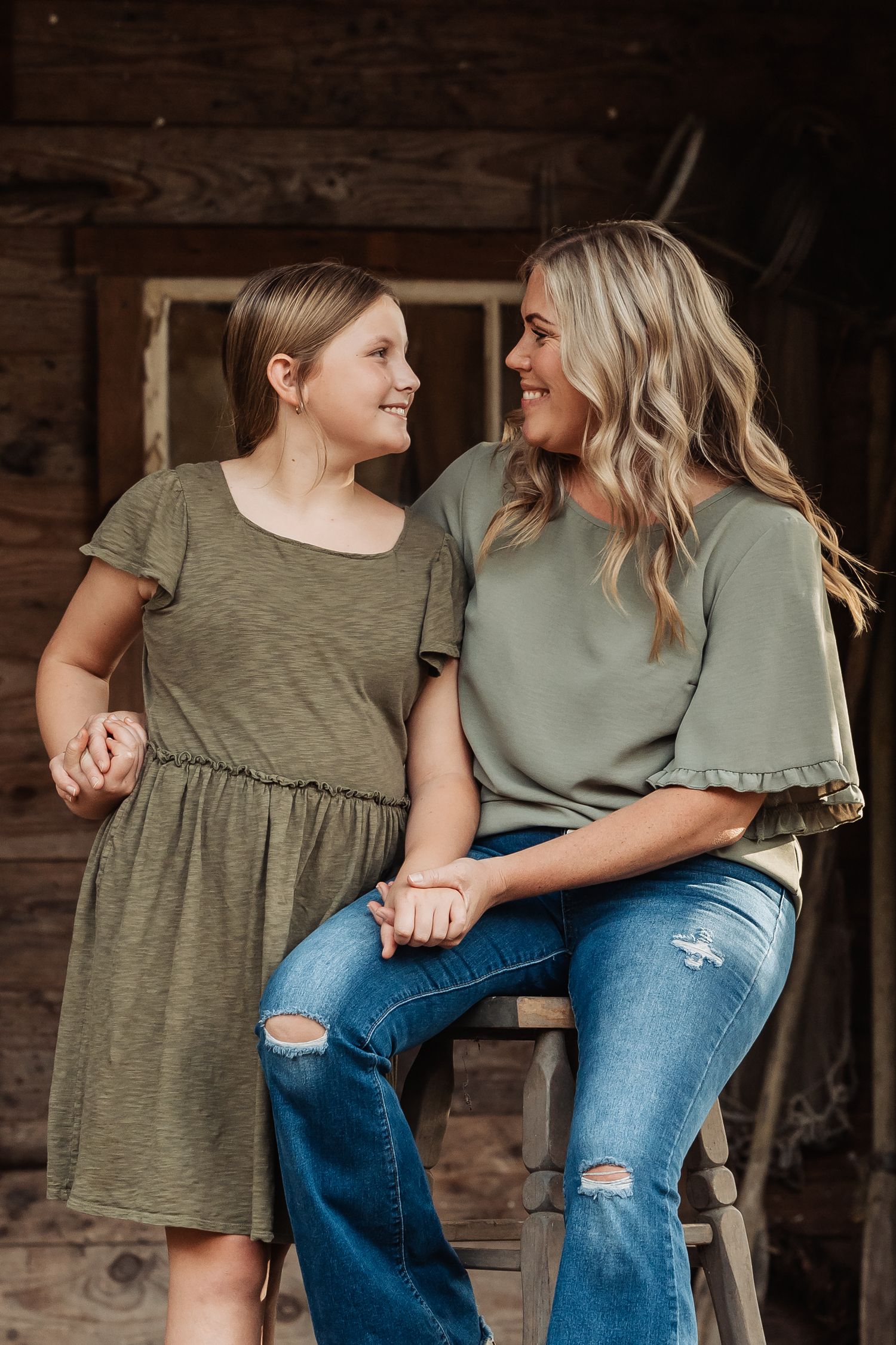 A mother and daughter in matching olive green outfits smile at each other while sitting on a wooden stool.