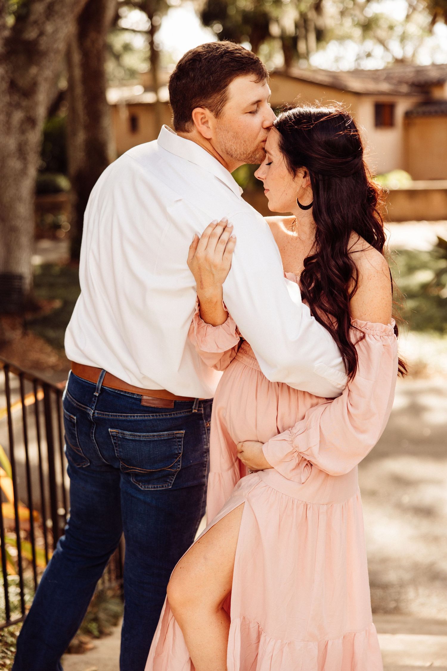 Couple embracing outdoors, pregnant woman in pink off-shoulder dress with partner.