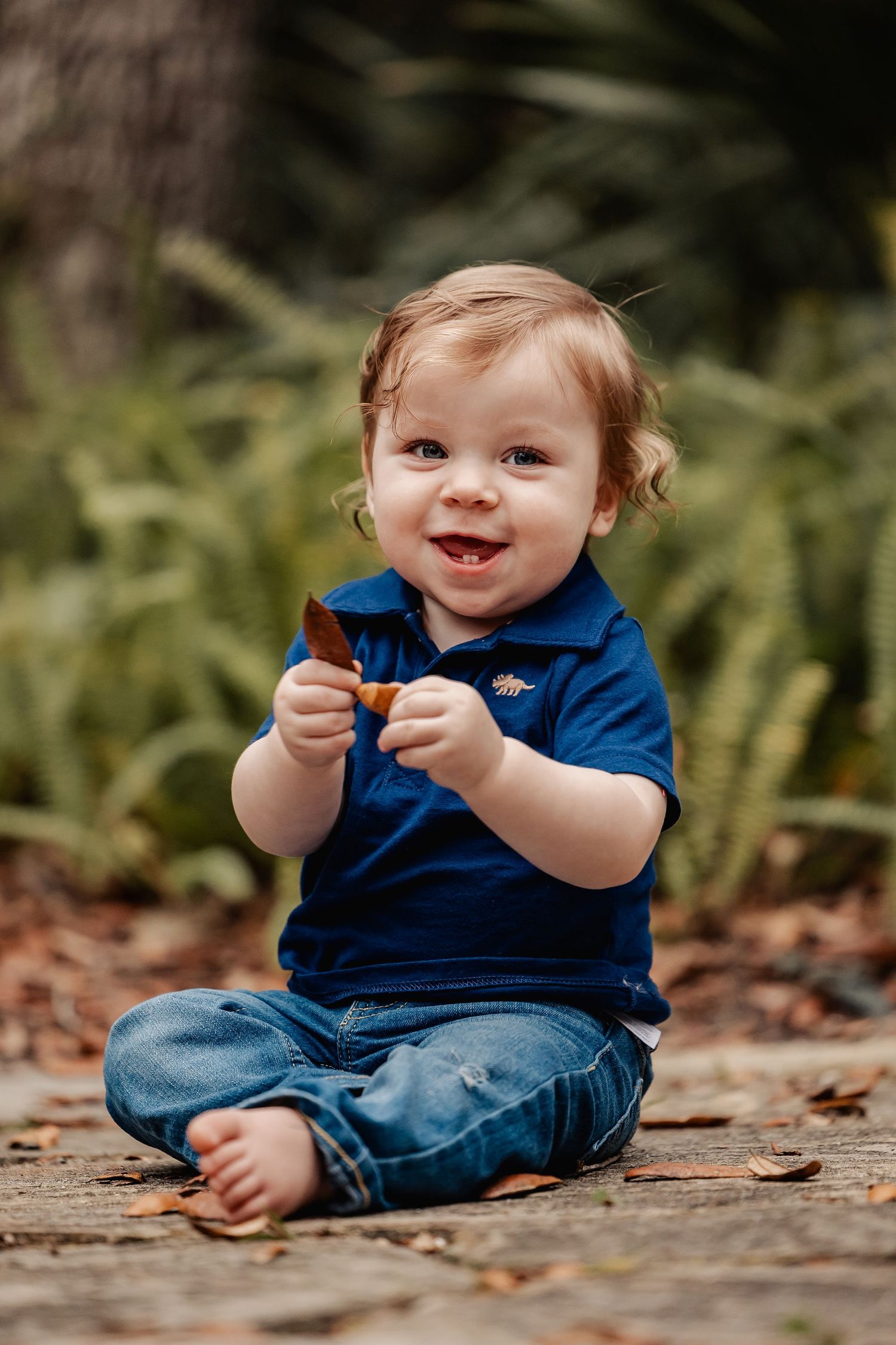 Adorable smiling baby boy in blue polo shirt and jeans sitting on ground holding a leaf outdoors.