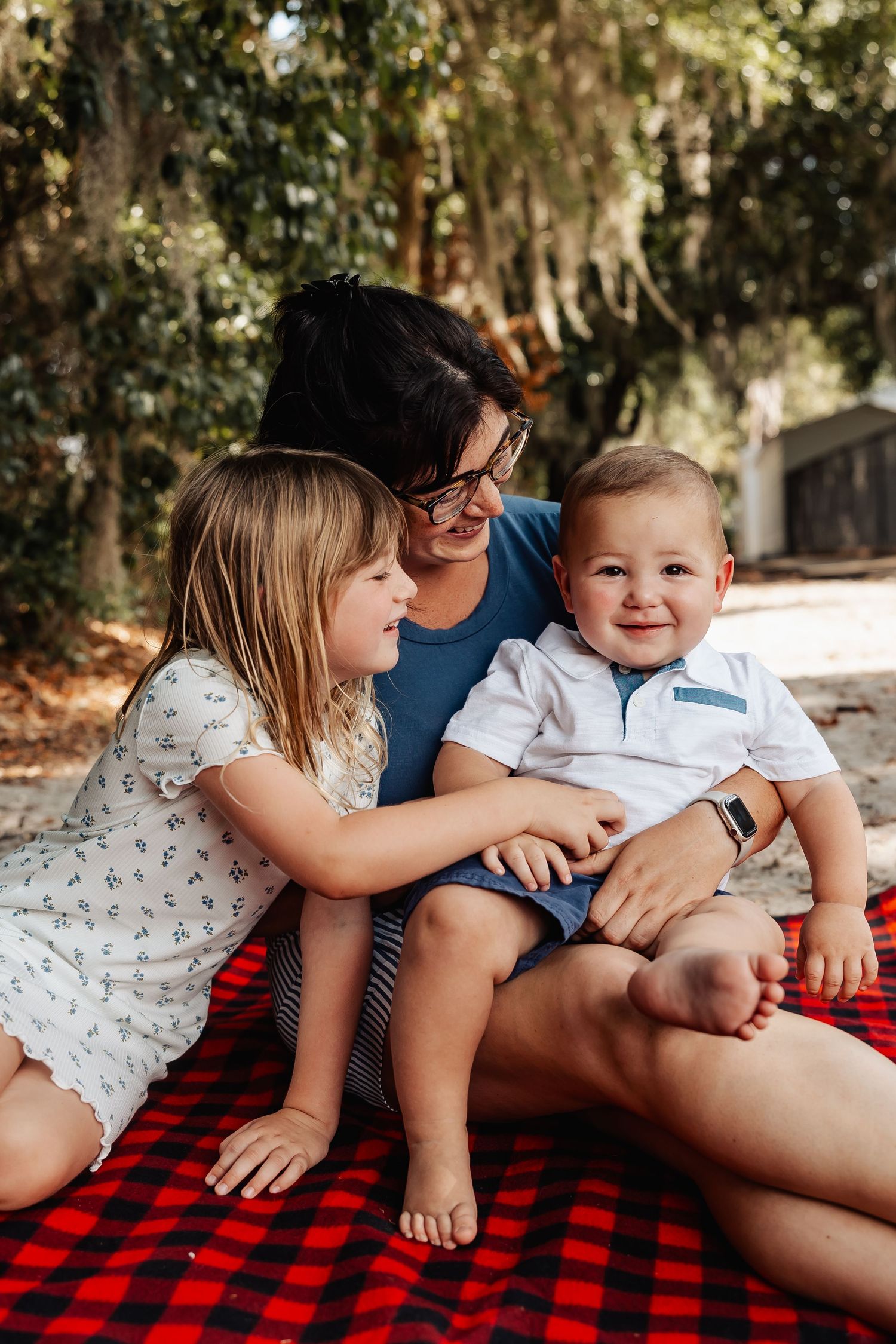 A grandmother sits on a red blanket outdoors with two young grandchildren smiling.