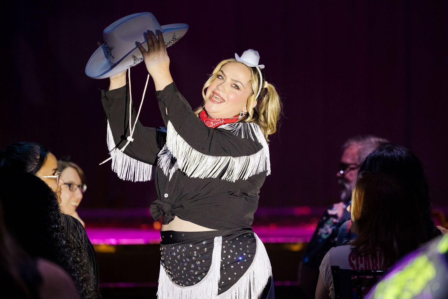 Female performer in black western fringe costume holding cowboy hat on stage.