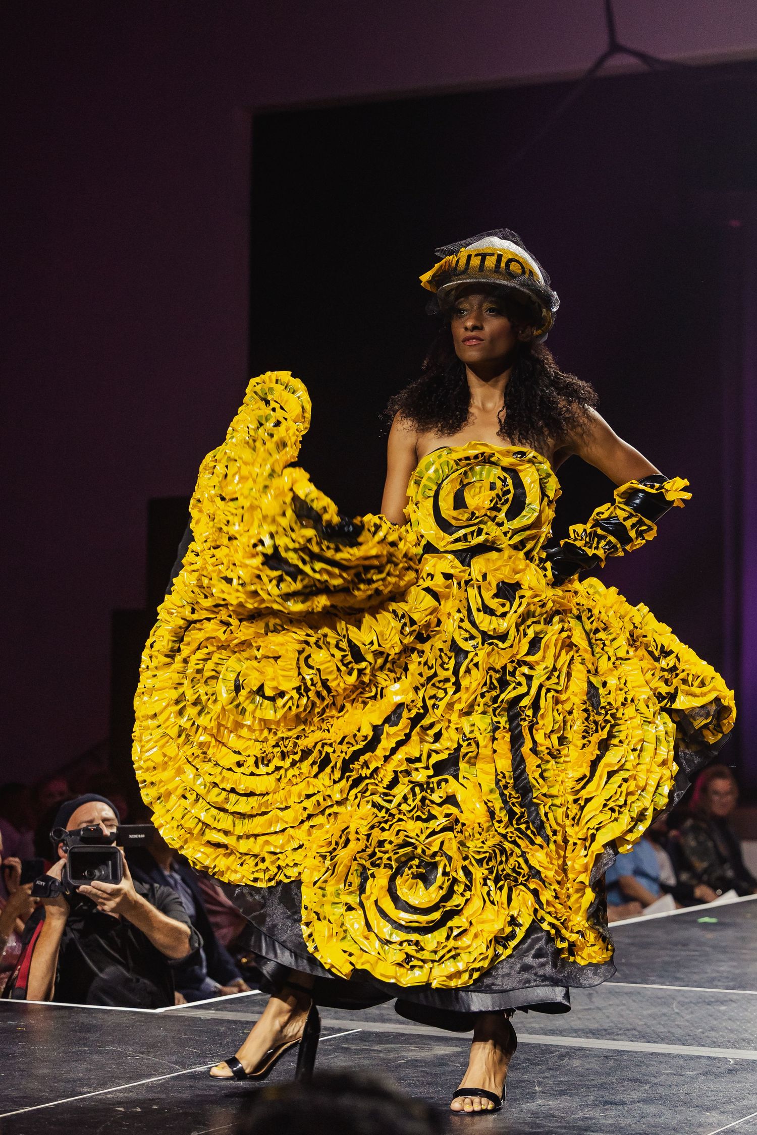 Model twirls in a stunning yellow ruffled balloon gown with matching hat on a fashion runway.