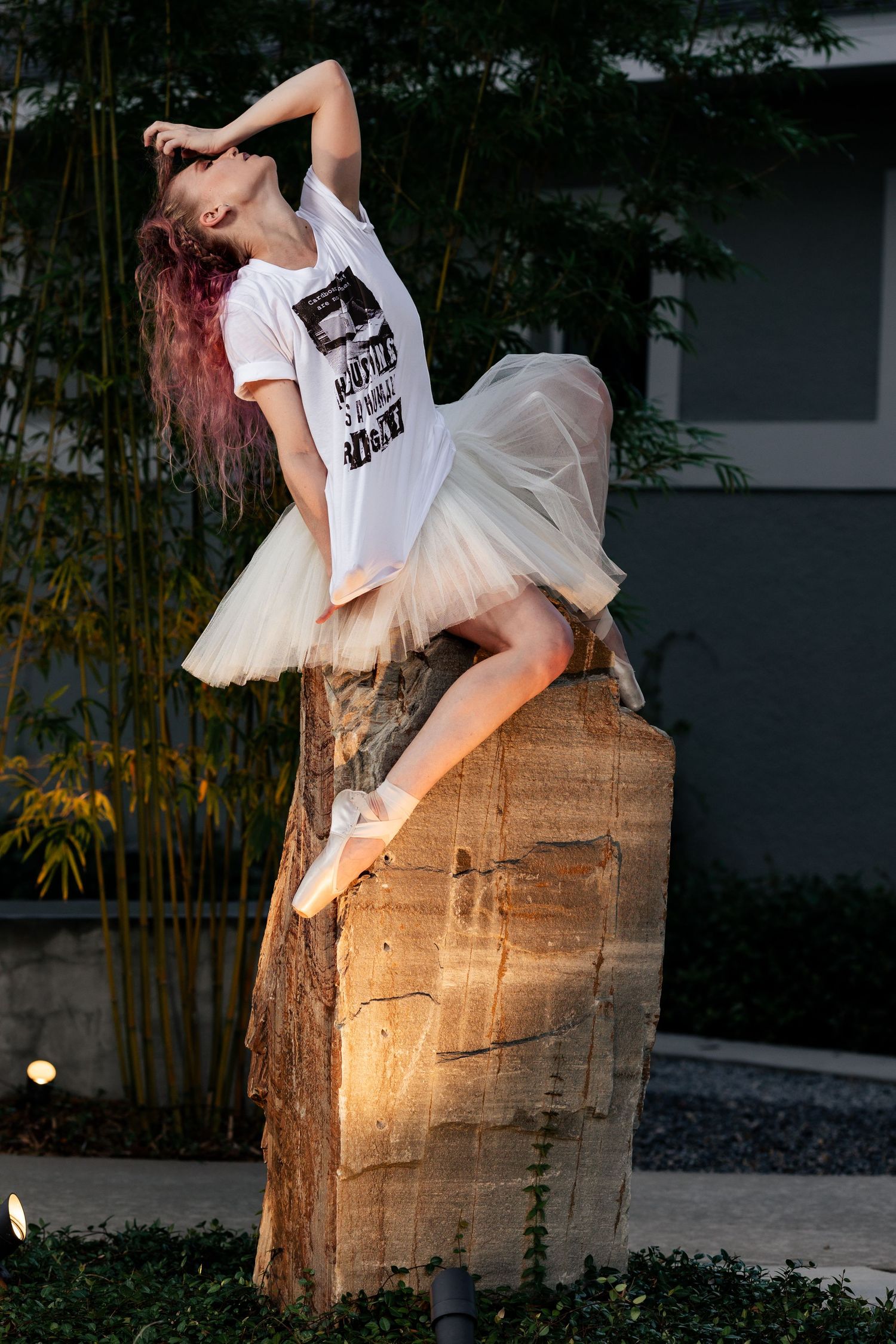 Red-haired ballerina in white tutu sitting gracefully on a stone pillar in an outdoor garden setting.