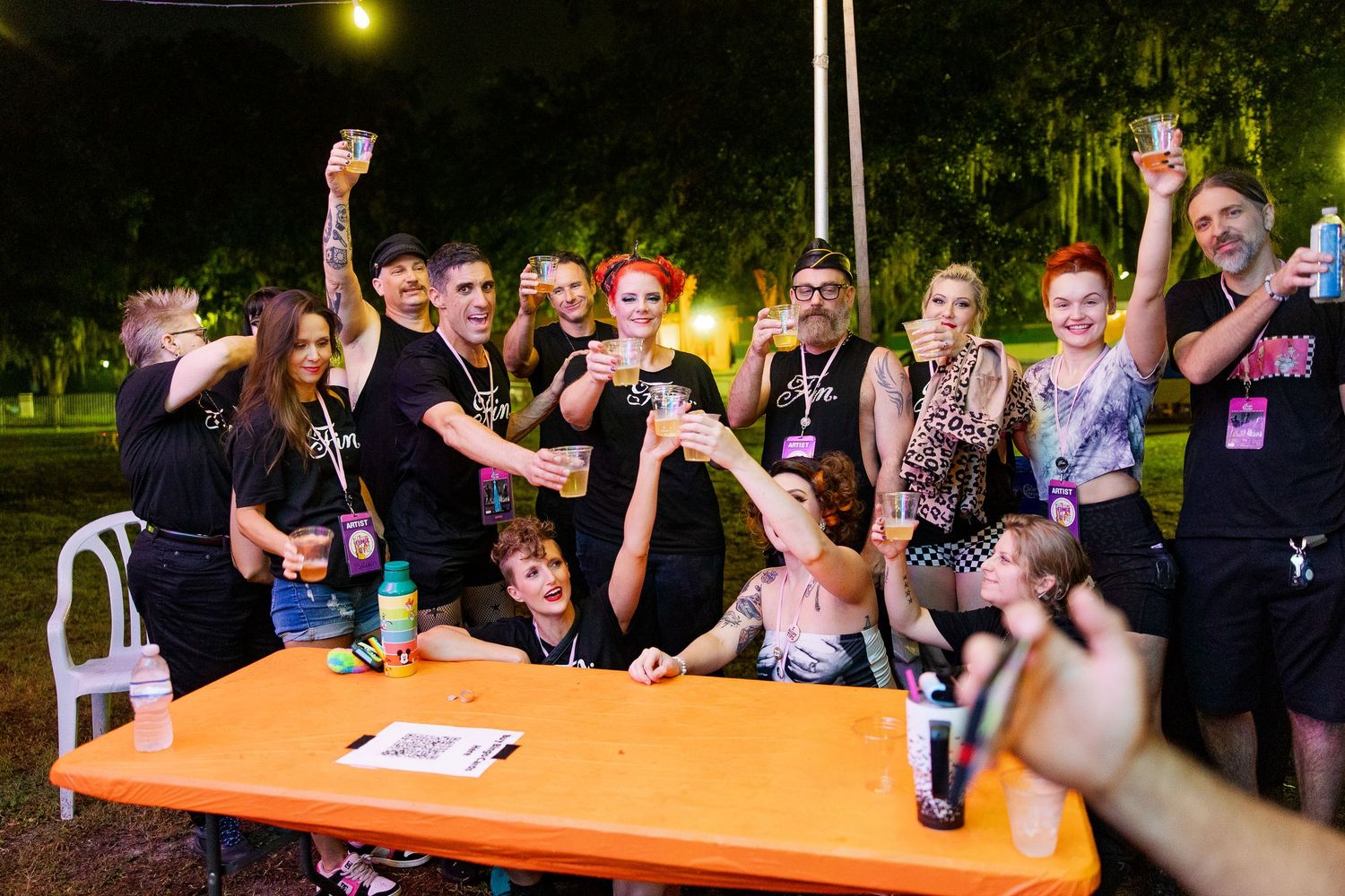 Group of friends raising drinks in a toast around an orange table at an outdoor night event.