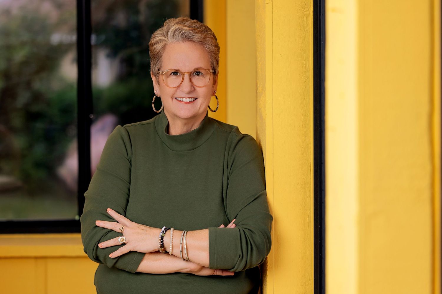 Woman with short gray hair in olive green sweater stands with arms crossed against a yellow wall outdoors.